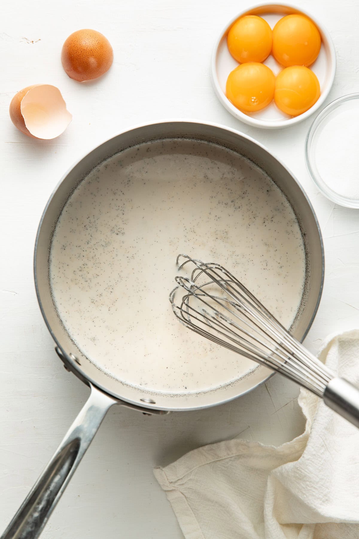 A saucepan filled with a creamy mixture is being whisked. Nearby are an empty eggshell, a plate with four egg yolks, and a small bowl of sugar on a white surface with a cloth.