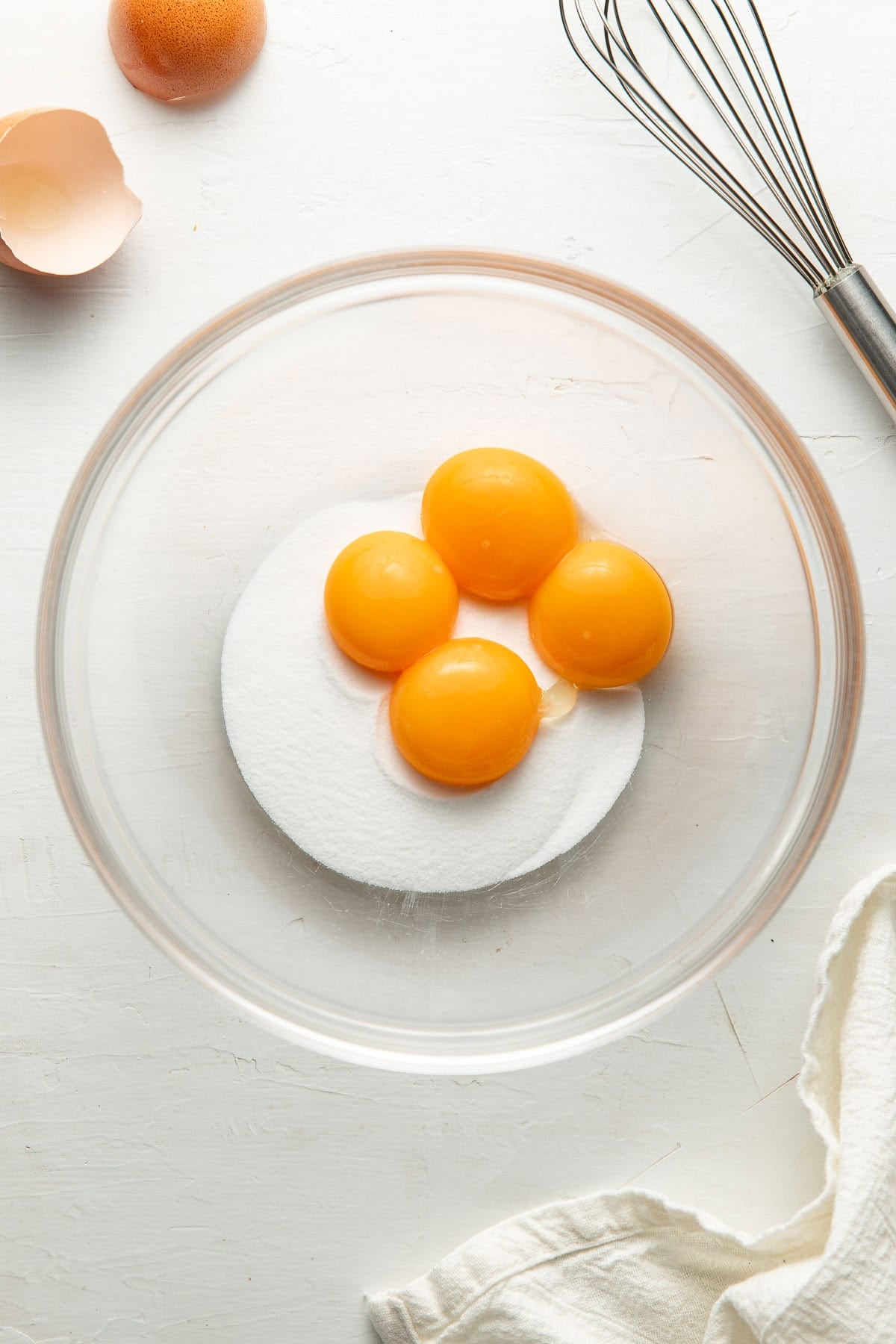 A clear glass bowl containing four egg yolks and white granulated sugar sits on a white surface near a cracked egg, a metal whisk, and a white cloth.