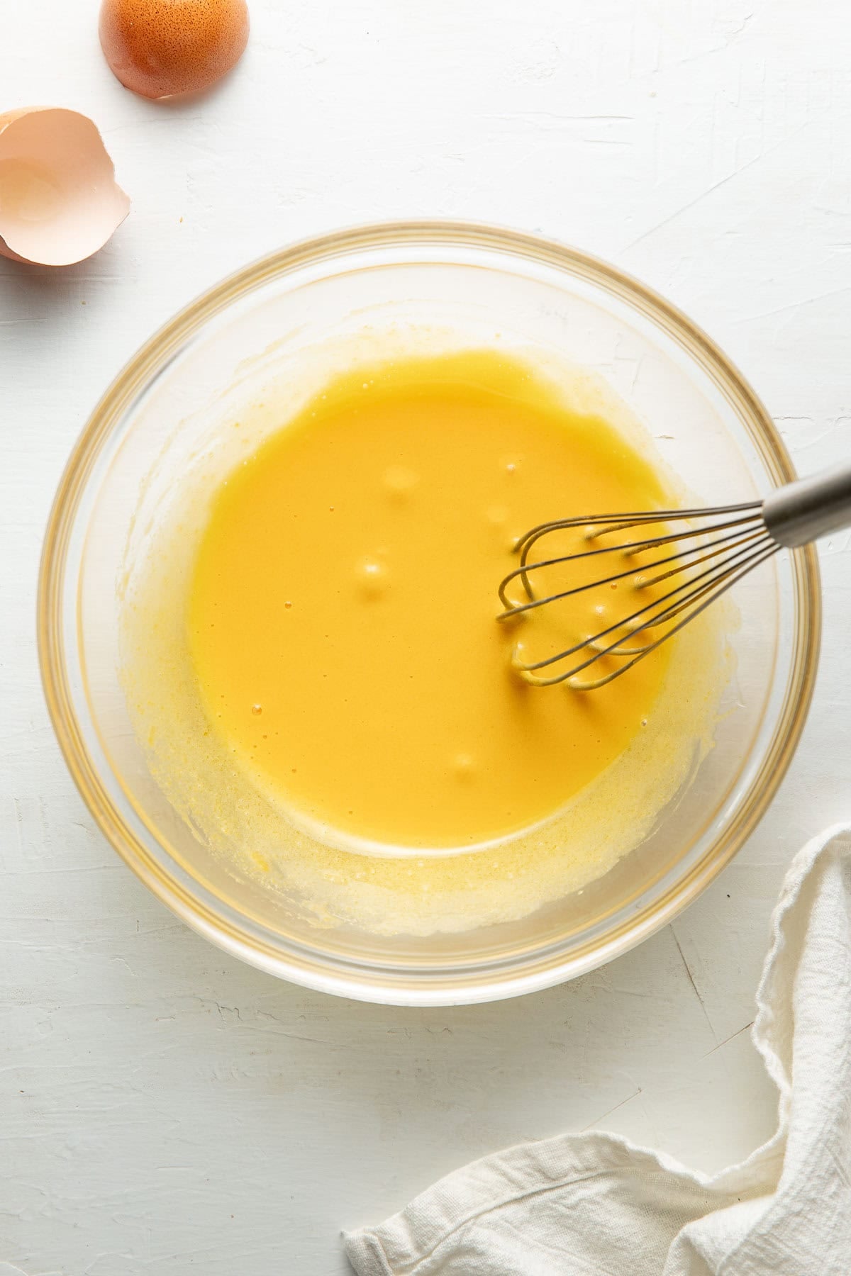 A glass bowl containing a yellow batter being whisked, with an egg shell and a whole egg nearby on a white surface, and a white cloth in the corner.
