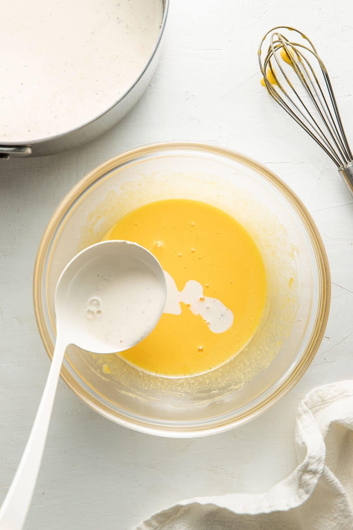 A ladle pours creamy liquid into a glass bowl with a yellow mixture. Nearby are a whisk with some yellow residue, a saucepan with more cream, and a light-colored cloth on a white surface.