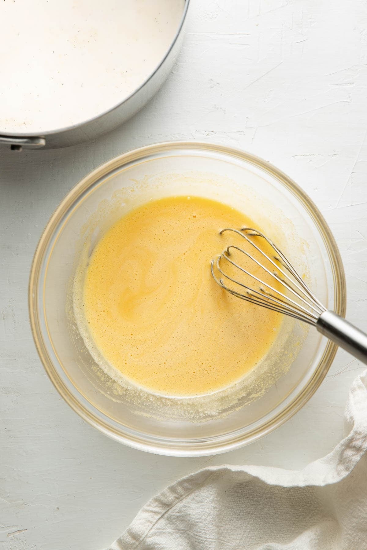 A glass bowl with a whisk inside contains a light yellow, frothy mixture. Nearby, part of a saucepan with a white liquid and a white cloth are visible on a light surface.