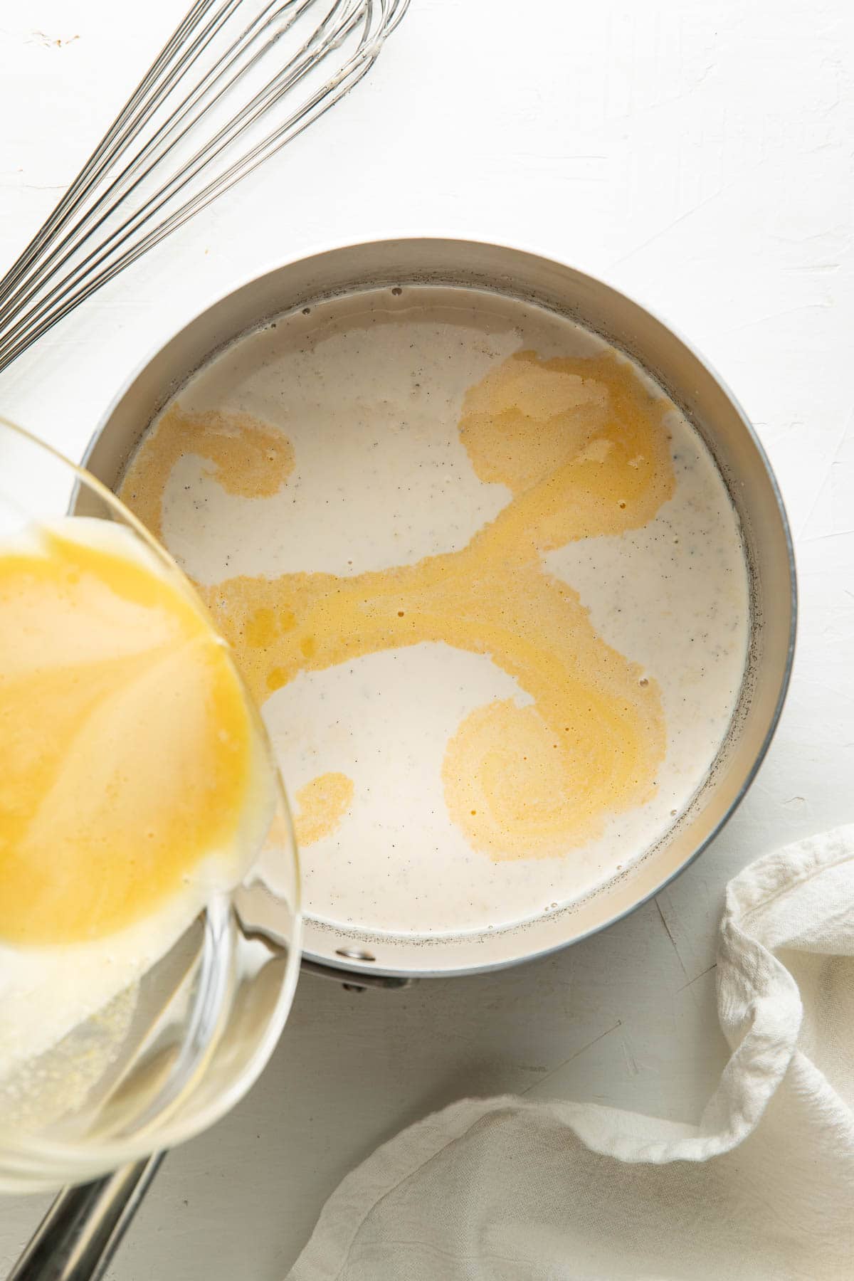 A saucepan filled with creamy liquid is on a white surface. Melted butter is being poured in, creating yellow swirls. A metal whisk and a white cloth are nearby.