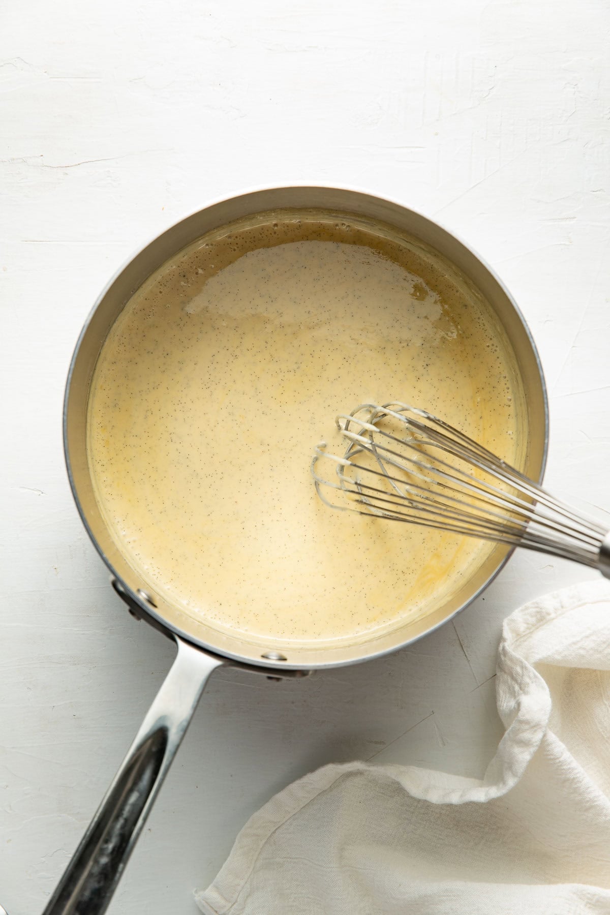 A saucepan filled with a creamy, light-yellow mixture is being whisked. The pan rests on a white surface beside a white cloth.