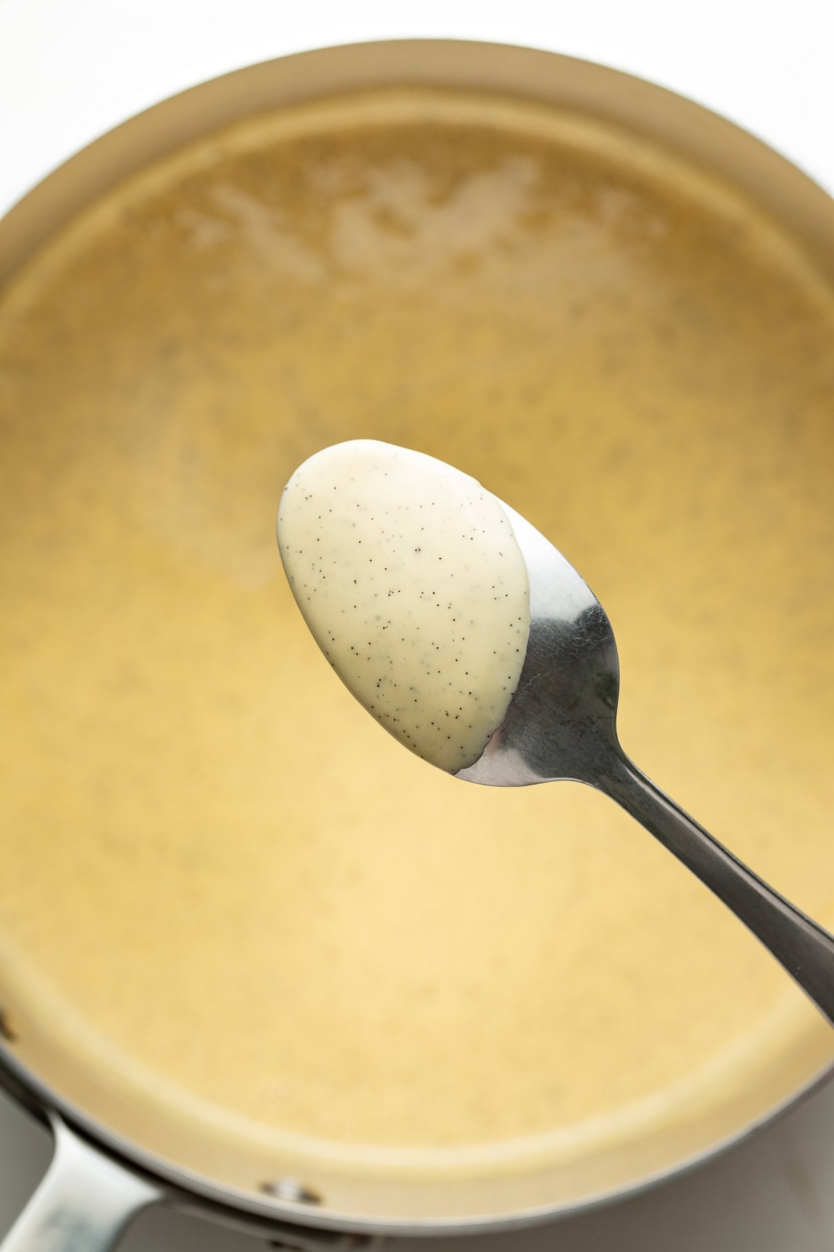 A spoon holding a thick, creamy sauce with visible black specks is held over a pan filled with the same sauce. The sauce appears smooth and light in color.