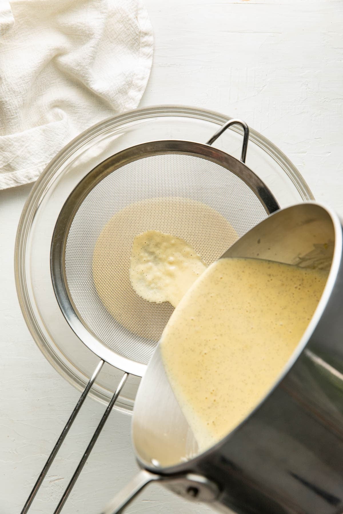 A creamy yellow mixture is being poured from a saucepan through a fine mesh strainer into a glass bowl on a white surface, with a white cloth nearby.