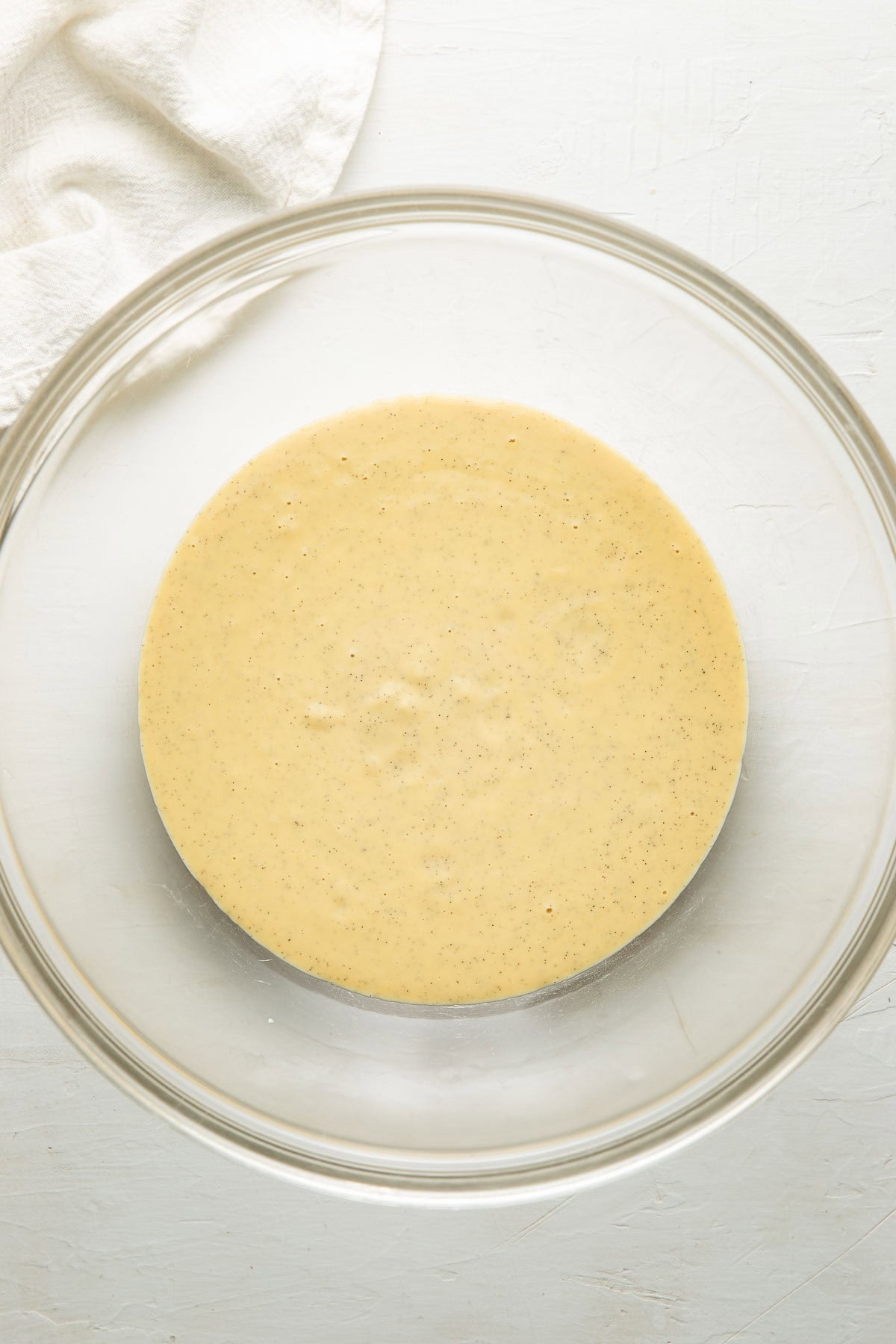 A glass mixing bowl filled with a smooth, pale yellow batter sits on a white surface, with part of a white cloth visible in the upper left corner.