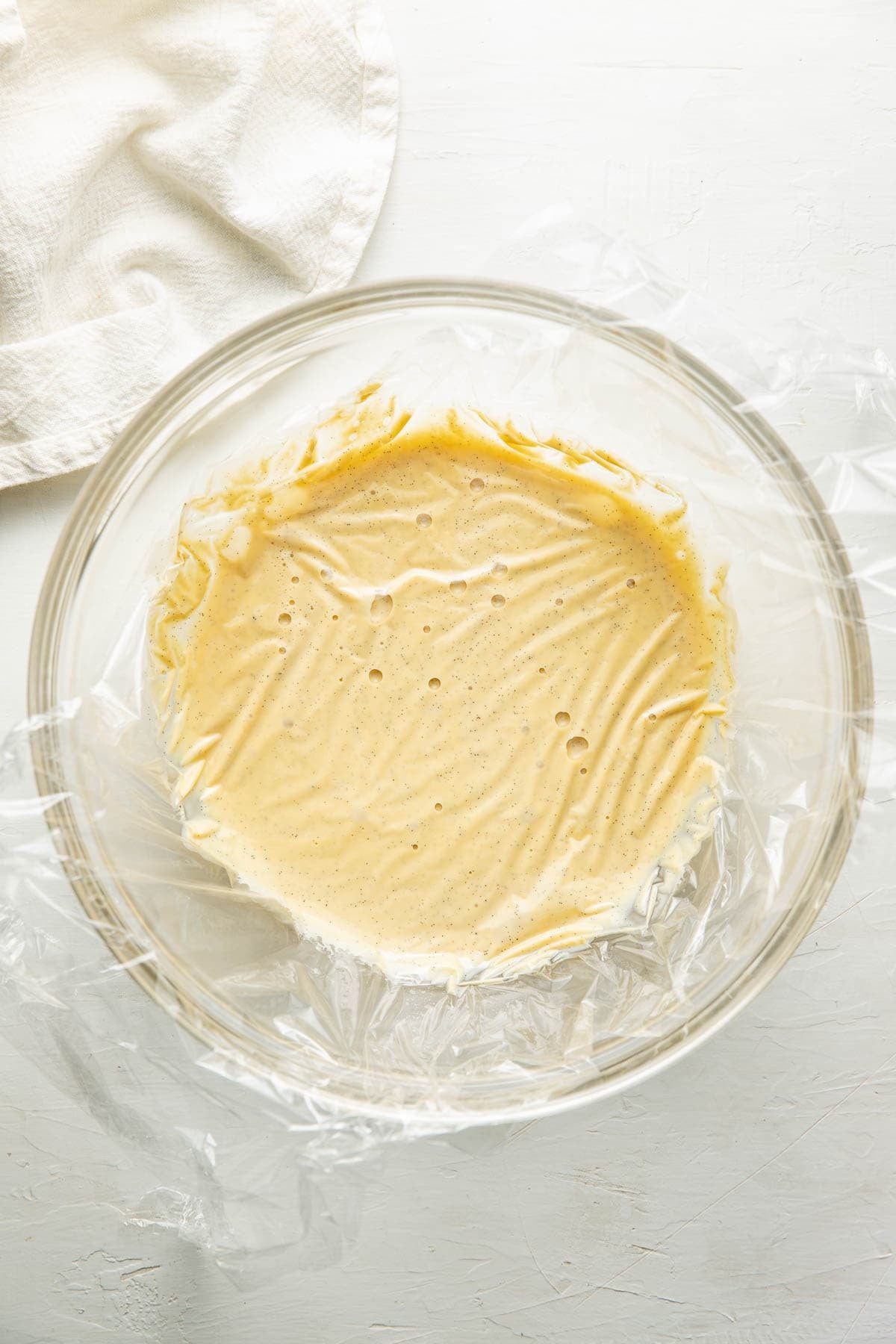 A glass bowl lined with plastic wrap contains pale yellow batter or dough. The bowl sits on a white surface next to a textured white cloth.