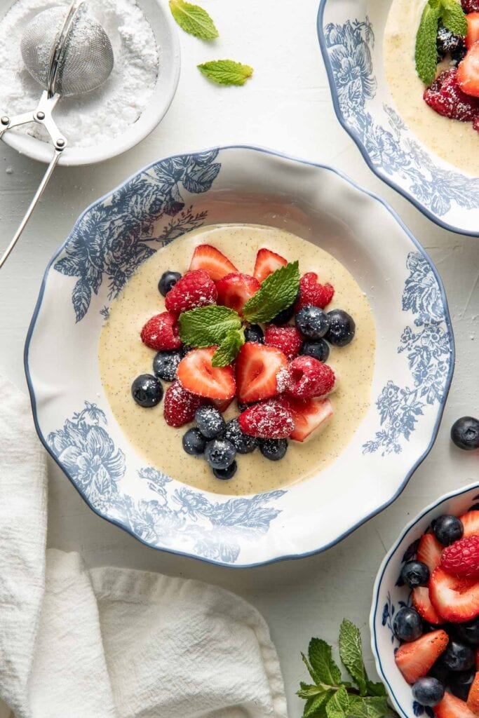 A blue and white floral bowl filled with berries—strawberries, raspberries, and blueberries—on a creamy sauce, garnished with mint and powdered sugar. Nearby are bowls, a sifter with powdered sugar, and a white napkin.