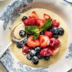 A bowl of creamy custard topped with fresh strawberries, raspberries, blueberries, a dusting of powdered sugar, and a sprig of mint; a spoon rests in the bowl which has a blue floral pattern.
