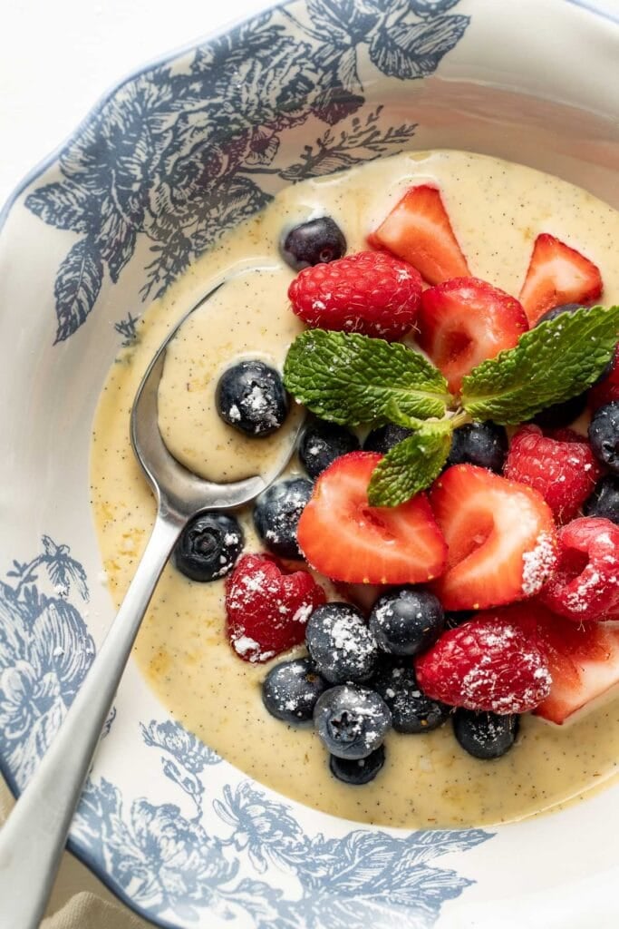 A bowl of creamy custard topped with fresh strawberries, blueberries, raspberries, a dusting of powdered sugar, and mint leaves, with a spoon resting inside the decorative bowl.