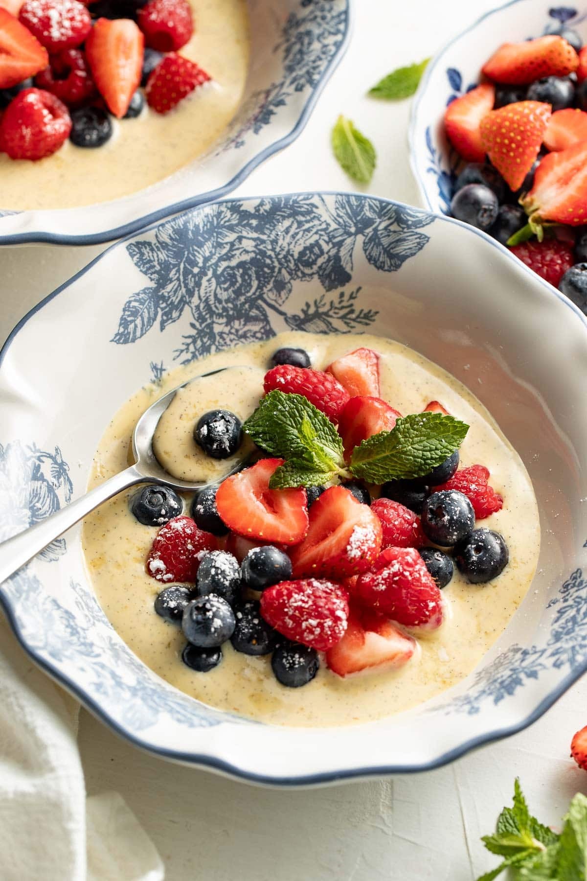 A bowl of creamy custard topped with fresh strawberries, blueberries, and raspberries, garnished with mint leaves. A spoon rests in the bowl, and two similar bowls are in the background.