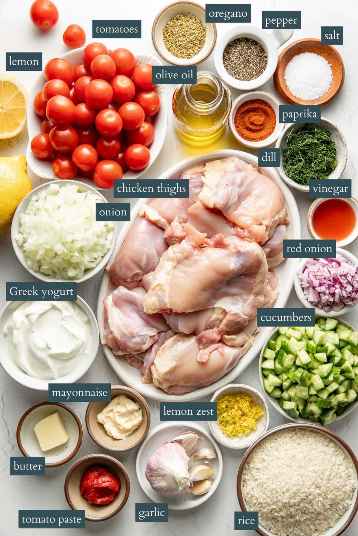 A flat lay of labeled ingredients for a recipe, including chicken thighs, tomatoes, onion, red onion, cucumbers, rice, Greek yogurt, mayonnaise, butter, tomato paste, lemon, lemon zest, garlic, olive oil, vinegar, dill, oregano, paprika, salt, and pepper.