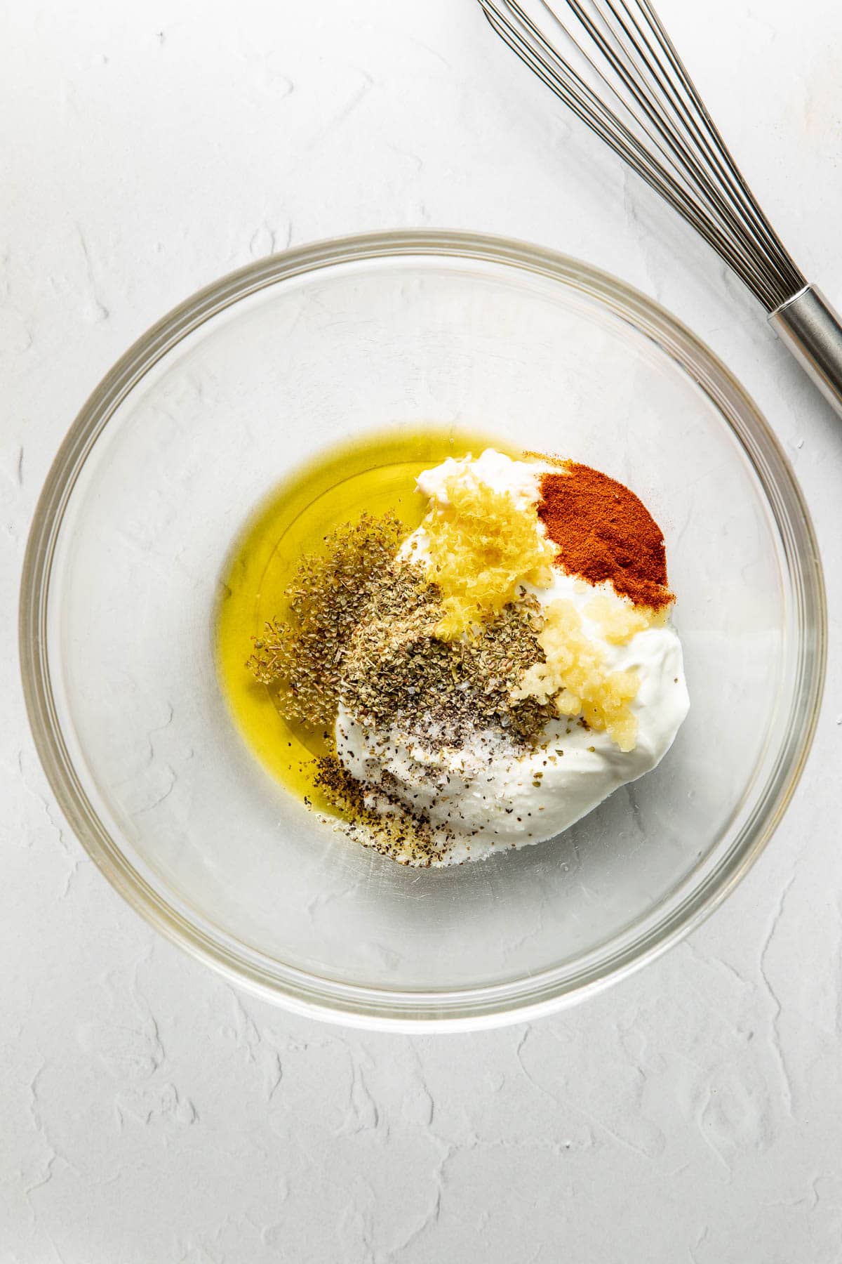 A glass bowl containing yogurt, olive oil, minced garlic, black pepper, paprika, and other seasonings sits on a white surface next to a metal whisk.