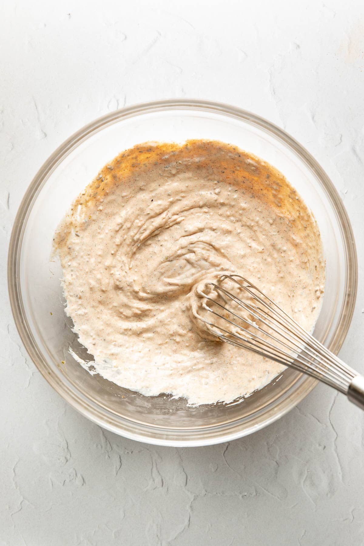 A glass bowl filled with a light brown batter being whisked on a white textured surface.