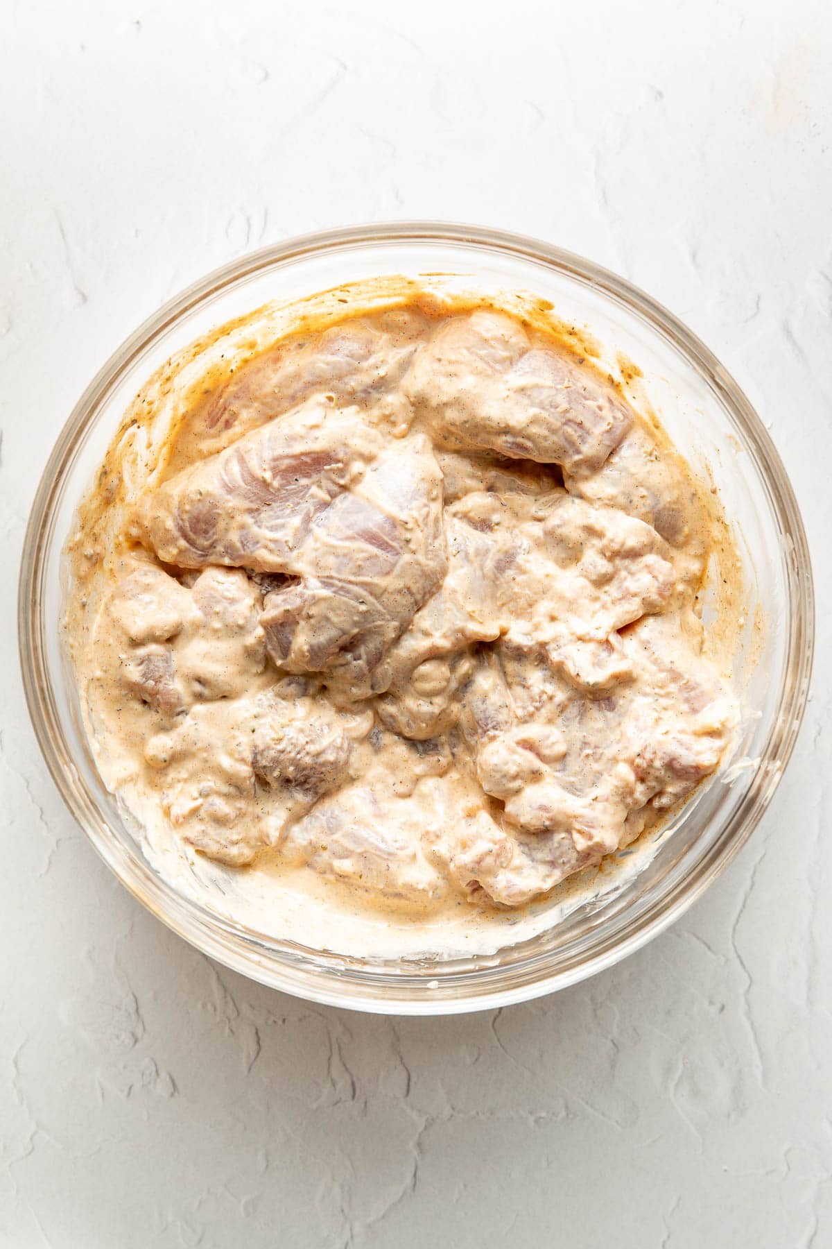 Raw chicken pieces coated in a creamy marinade are placed in a clear glass bowl on a white textured surface.