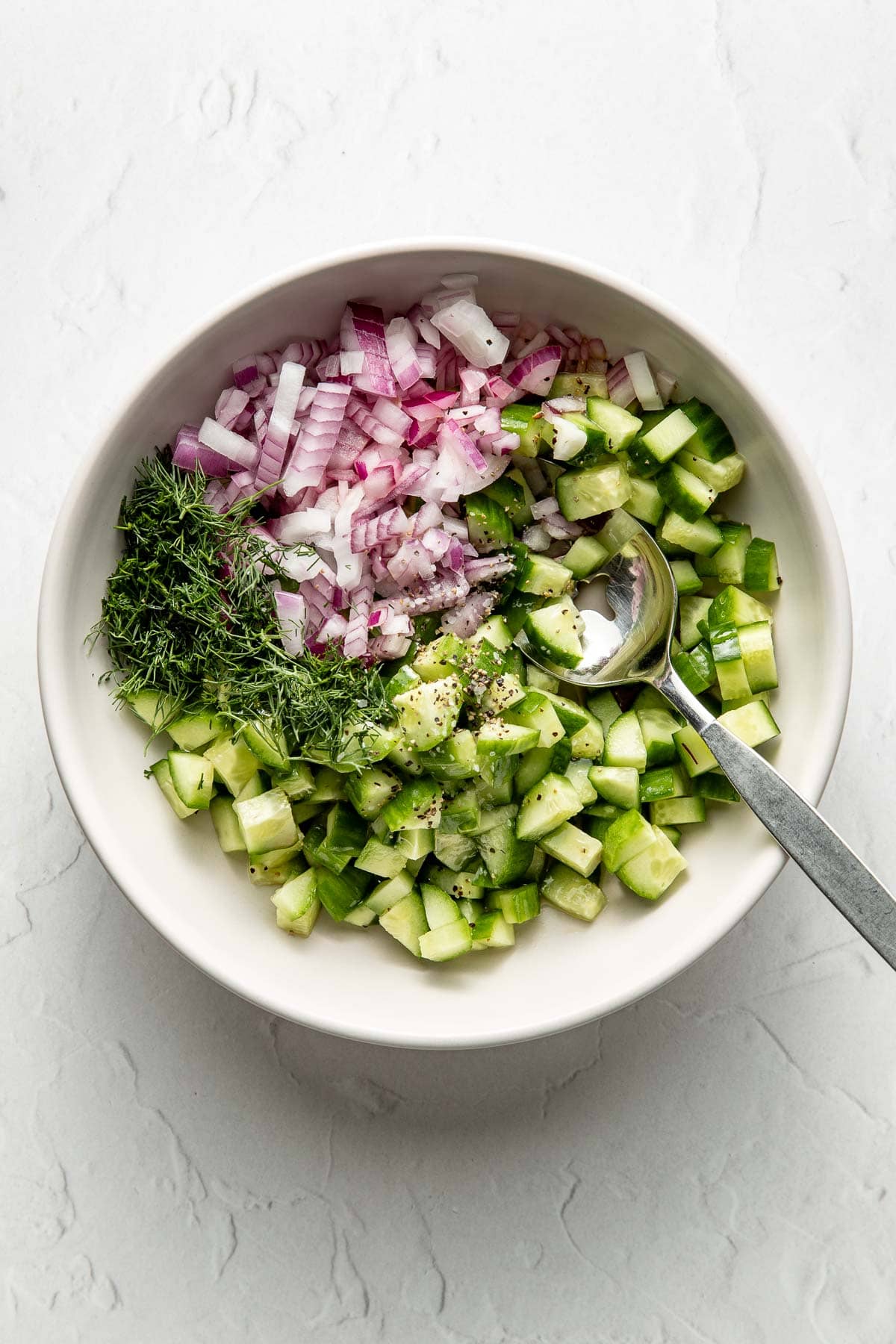 A white bowl filled with chopped red onions, cucumbers, and fresh dill, with black pepper sprinkled on top and a metal spoon resting inside the bowl. The bowl sits on a light, textured surface.