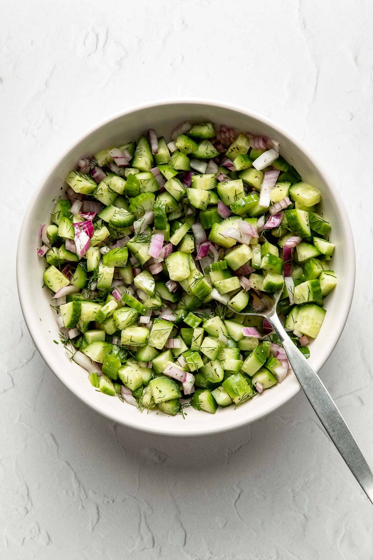 A white bowl filled with a chopped cucumber, green bell pepper, and red onion salad, garnished with herbs, on a light marble surface. A silver spoon rests inside the bowl.