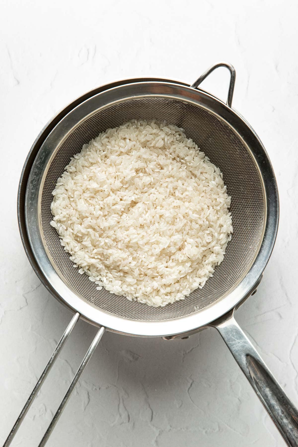 A metal strainer filled with uncooked white rice is placed over a stainless steel bowl on a white surface.