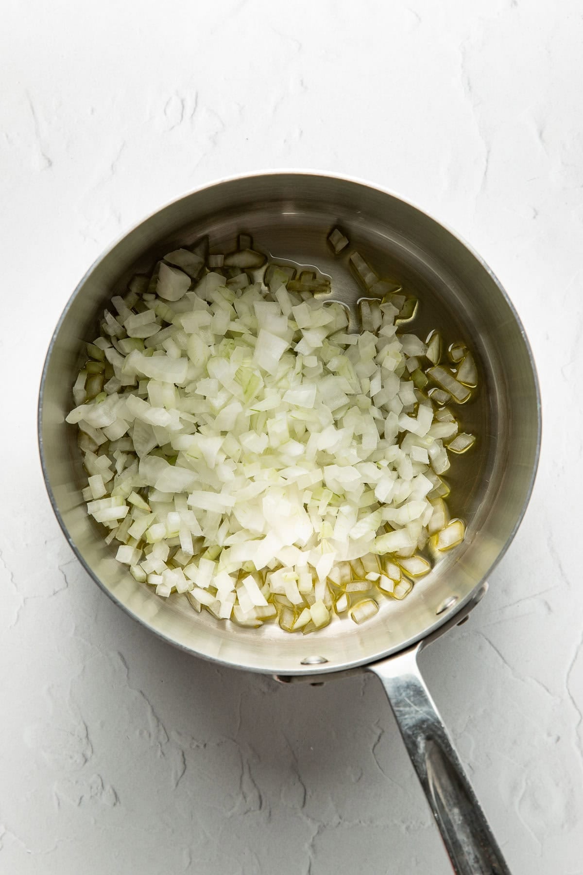 A stainless steel saucepan containing chopped onions and a small amount of olive oil, placed on a white textured surface.