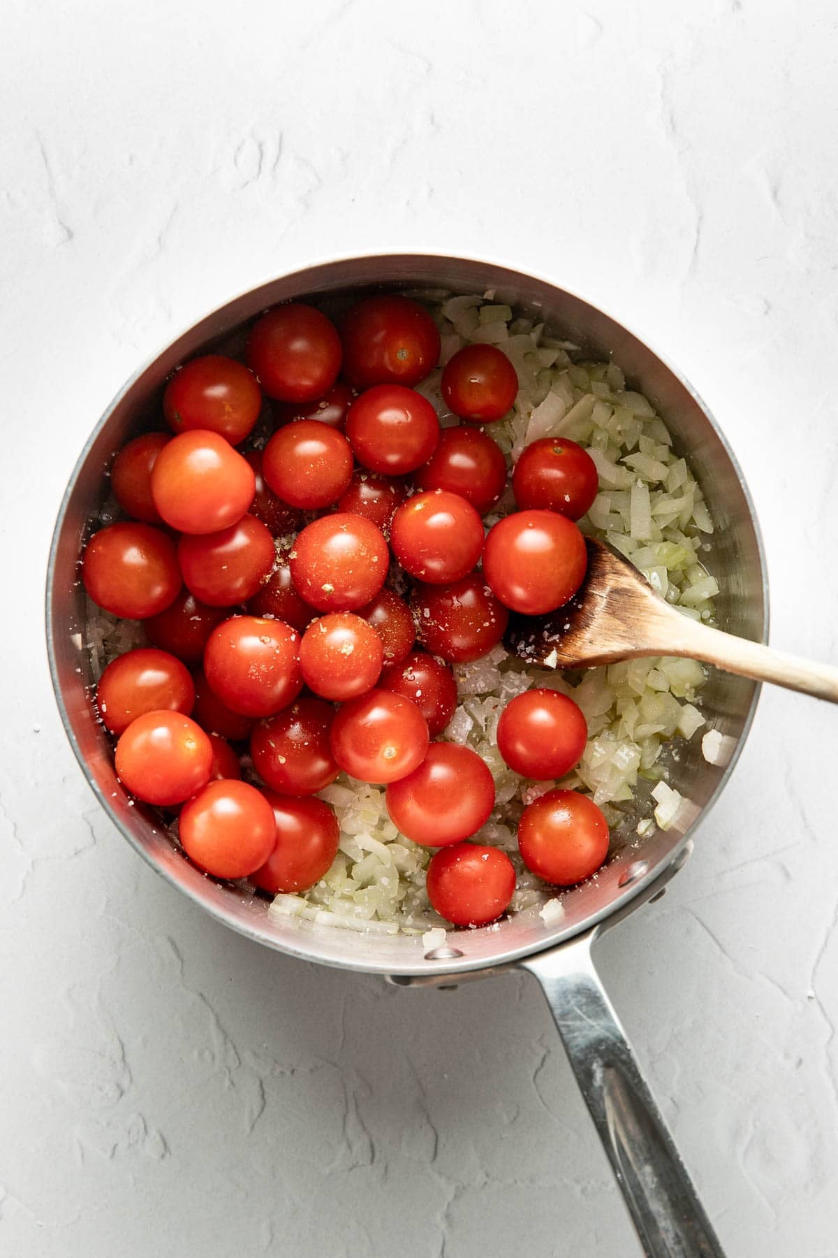 A stainless steel saucepan filled with chopped onions and whole cherry tomatoes sits on a light surface. A wooden spoon rests in the pan, ready to stir the ingredients.