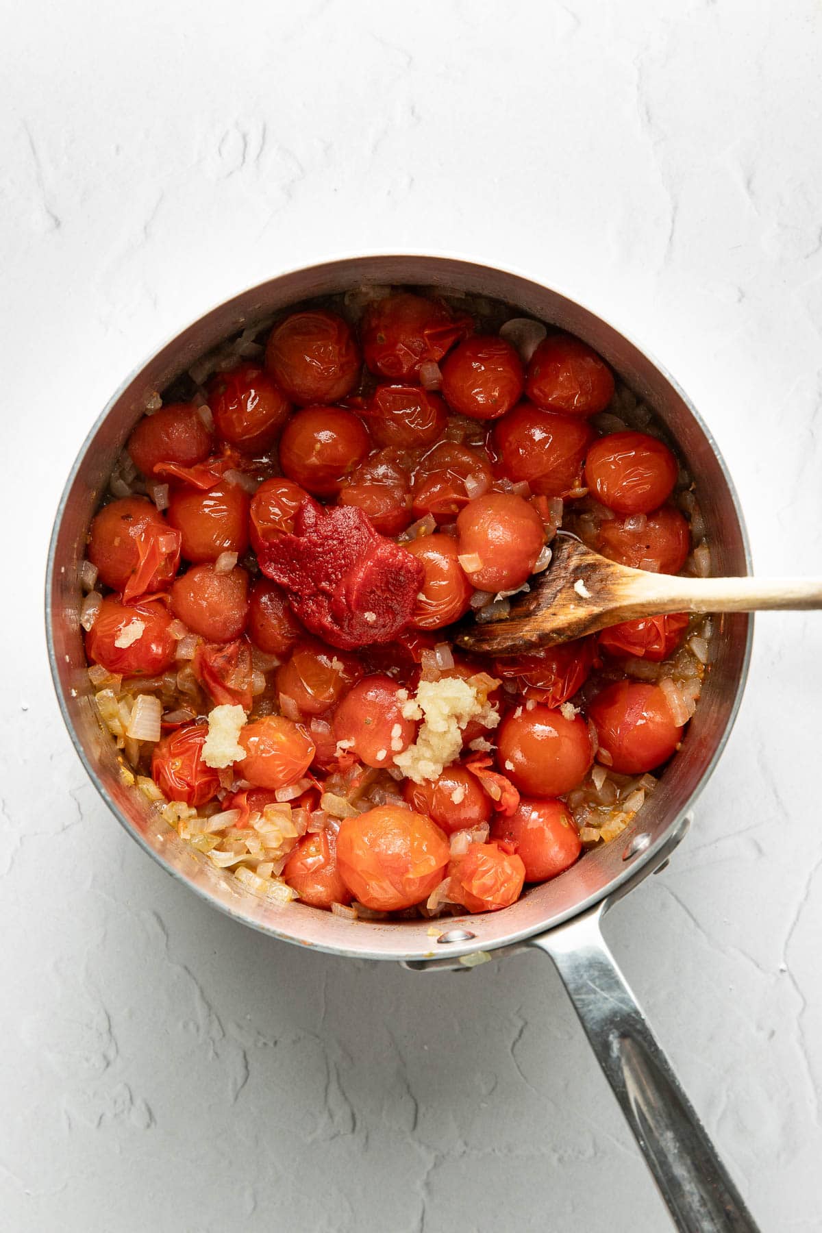 A saucepan filled with cherry tomatoes, chopped onions, tomato paste, and minced garlic being stirred with a wooden spoon on a white textured surface.