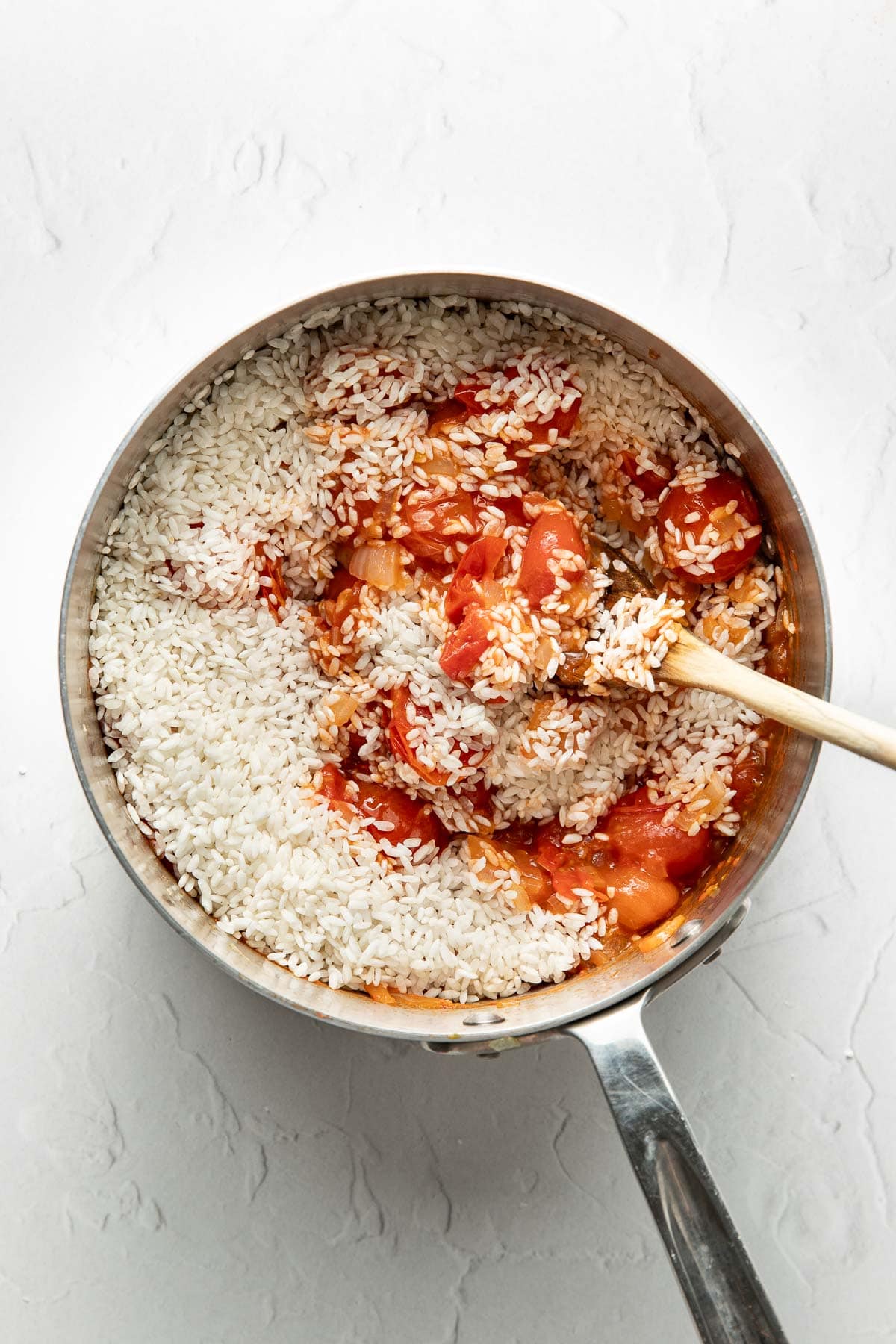 A stainless steel pot filled with uncooked white rice, chopped tomatoes, and onions being stirred with a wooden spoon on a white textured surface.