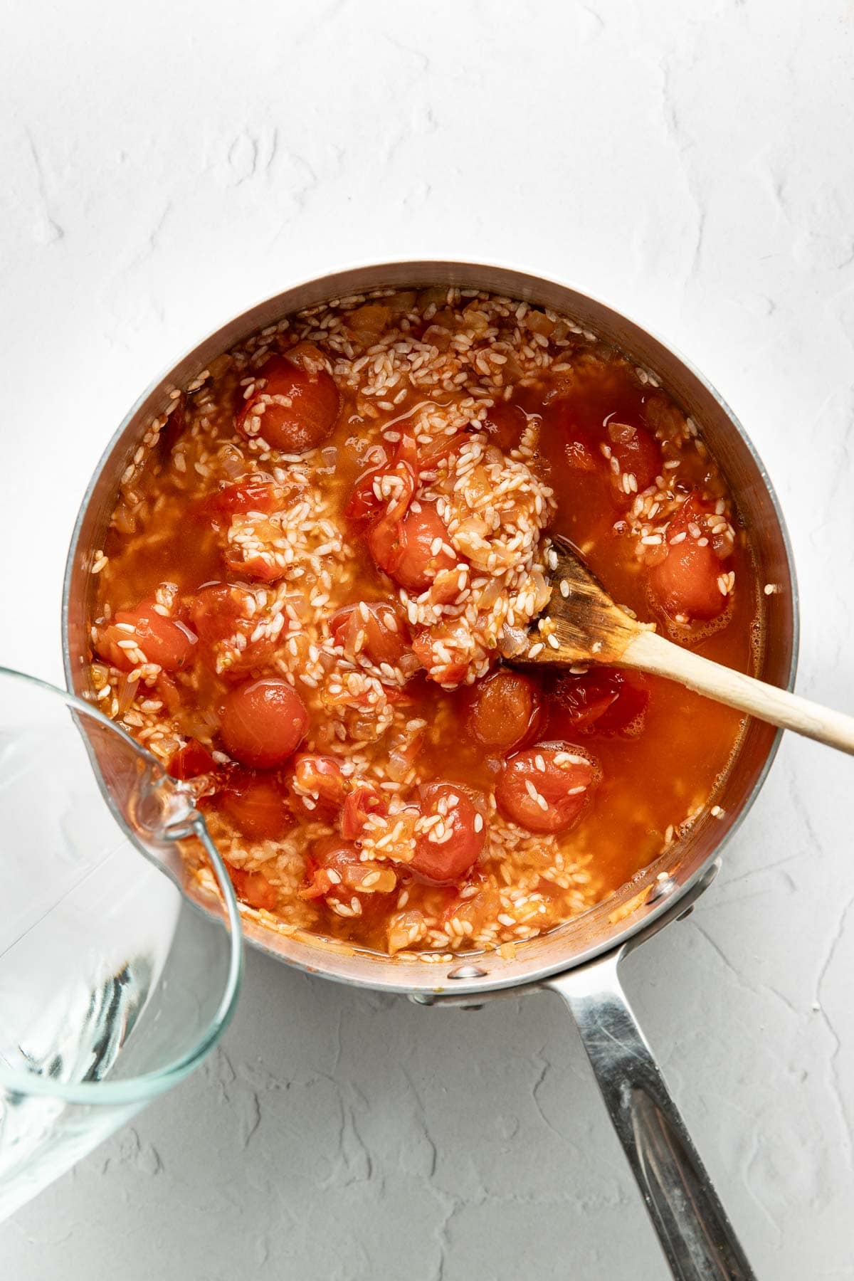 A saucepan filled with rice, whole cherry tomatoes, and liquid, being stirred with a wooden spoon. A glass container is pouring water into the pot. The scene is set on a white textured surface.