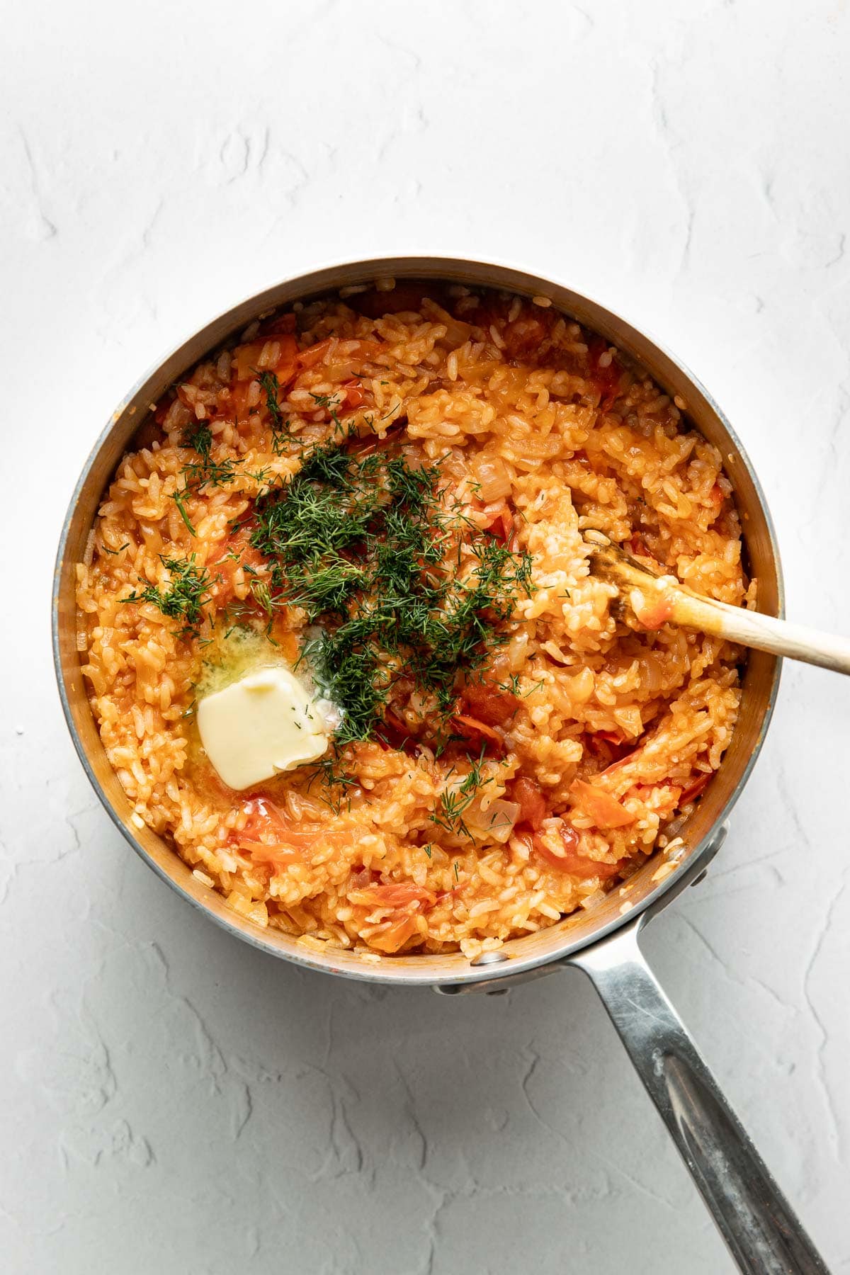 A saucepan filled with cooked tomato rice, topped with a pat of melting butter and chopped fresh herbs, sits on a white textured surface. A wooden spoon rests inside the pot.