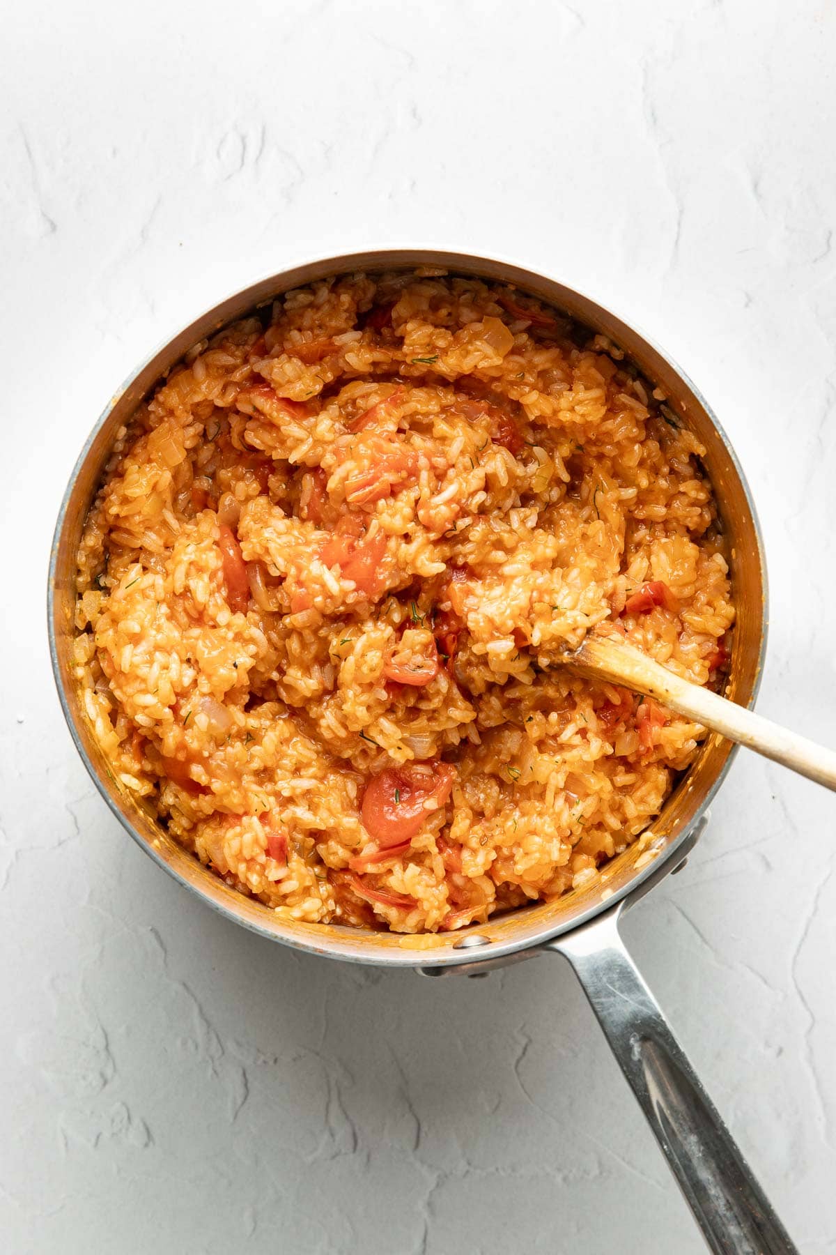 A stainless steel saucepan filled with creamy, orange-colored tomato rice, stirred with a wooden spoon, sits on a white textured surface.