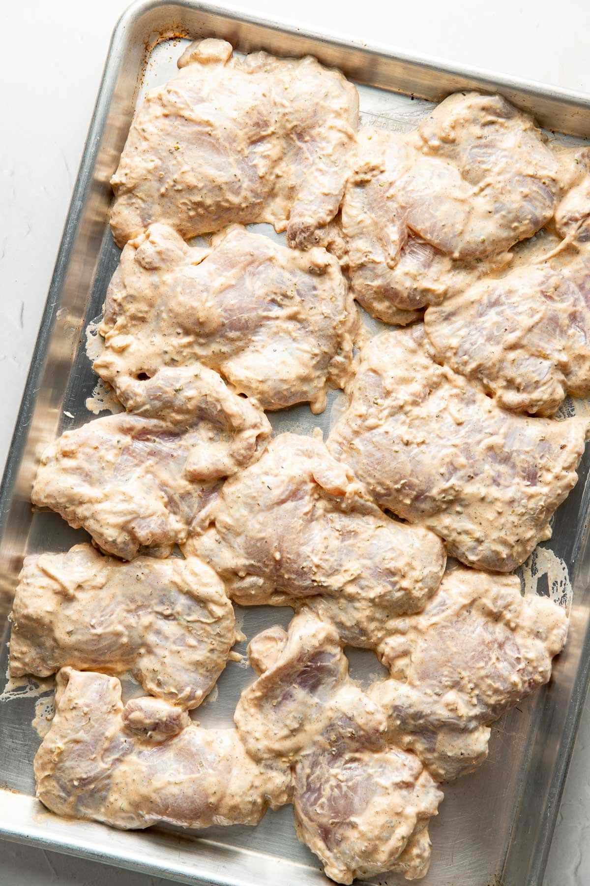 Raw chicken pieces coated in a creamy, light brown marinade are spread out on a metal baking sheet, ready to be cooked.