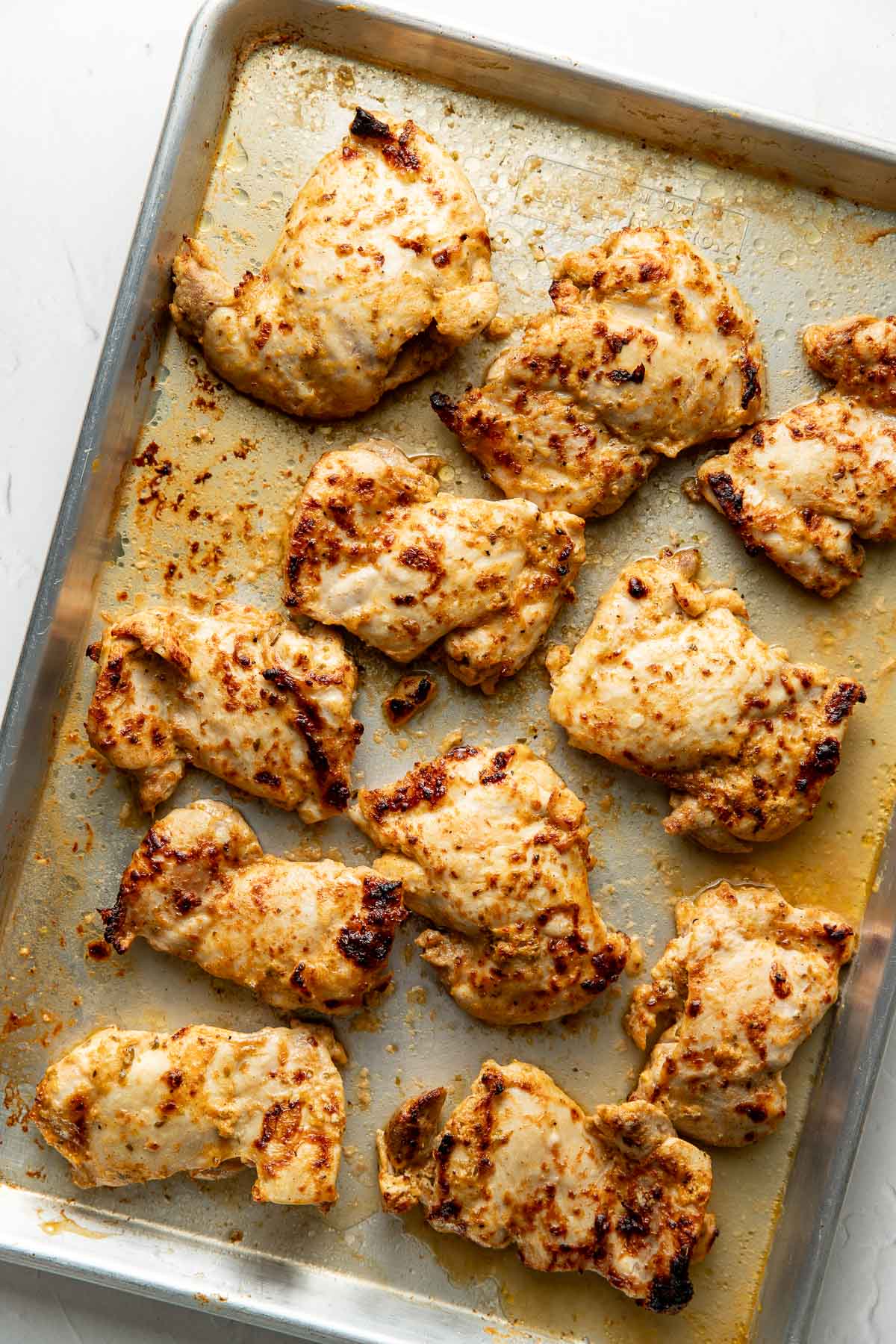 Baked, marinated chicken thighs arranged on a metal baking sheet, showing golden-brown, slightly charred edges and a juicy texture.