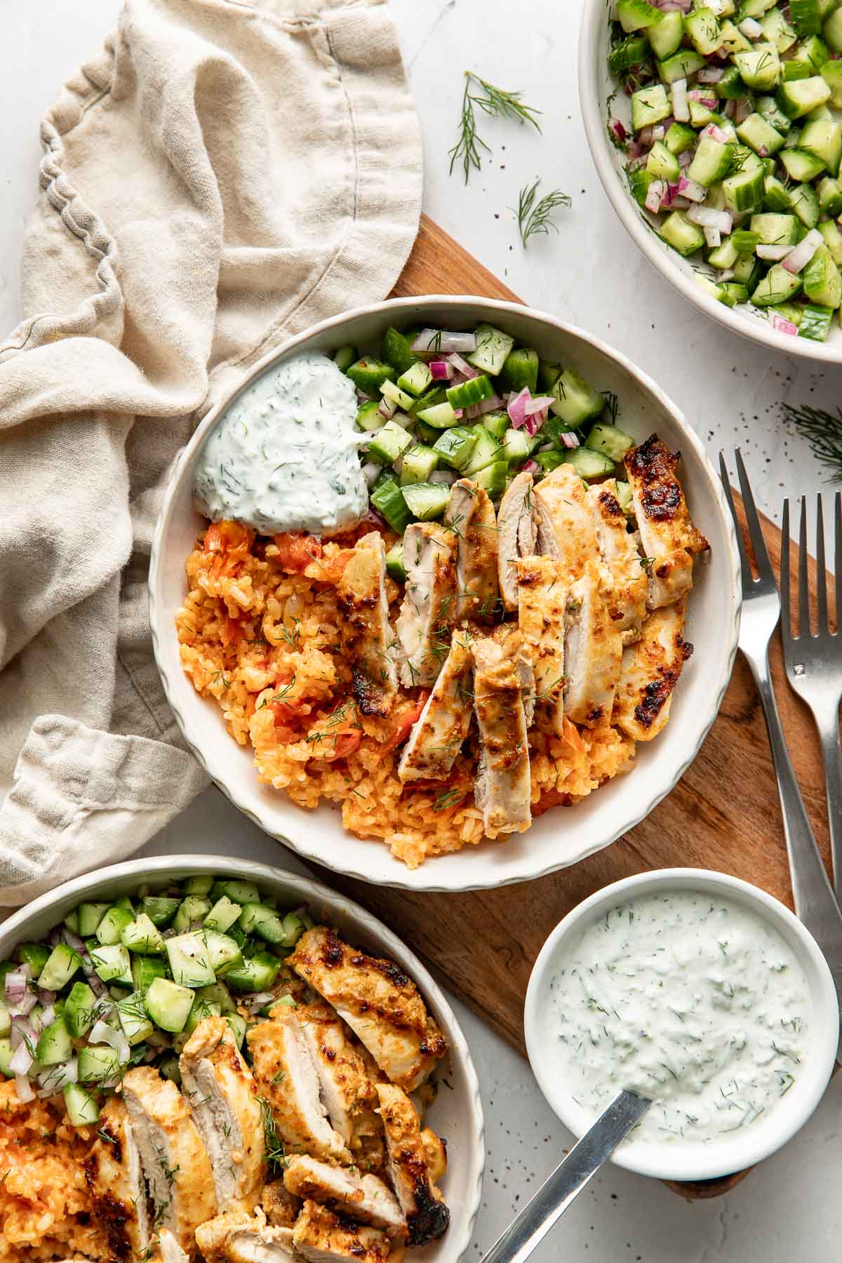 Two bowls filled with grilled chicken slices, yellow rice, chopped cucumber salad, and a dollop of creamy white sauce, arranged on a table with a beige napkin, extra sauce, and forks nearby.