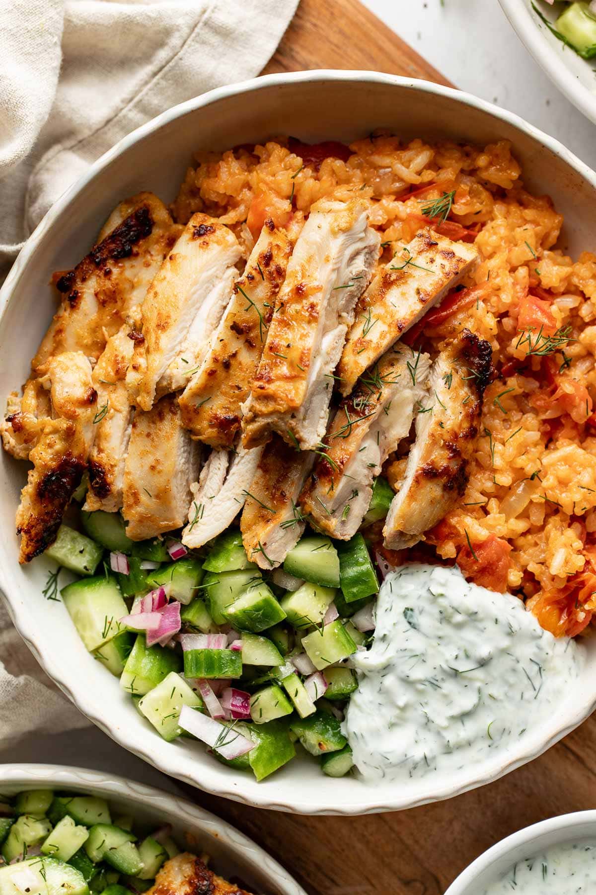 A bowl filled with sliced grilled chicken on seasoned rice, alongside a fresh cucumber and red onion salad, and a dollop of creamy herb sauce.