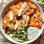 A bowl filled with grilled chicken slices, tomato rice, chopped cucumber salad with red onion, and a dollop of tzatziki sauce, all arranged neatly with a fork resting in the bowl.