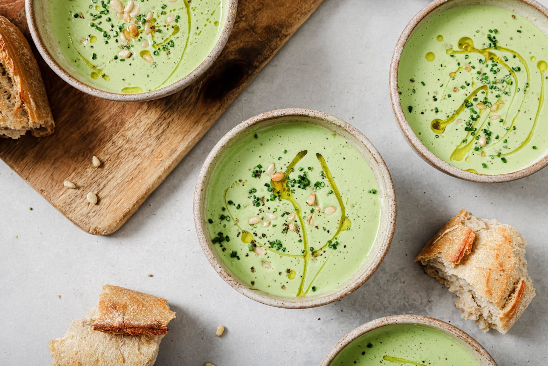 Three bowls of creamy green soup garnished with herbs, seeds, and a drizzle of olive oil sit on a light surface, surrounded by pieces of rustic bread and a wooden board.
