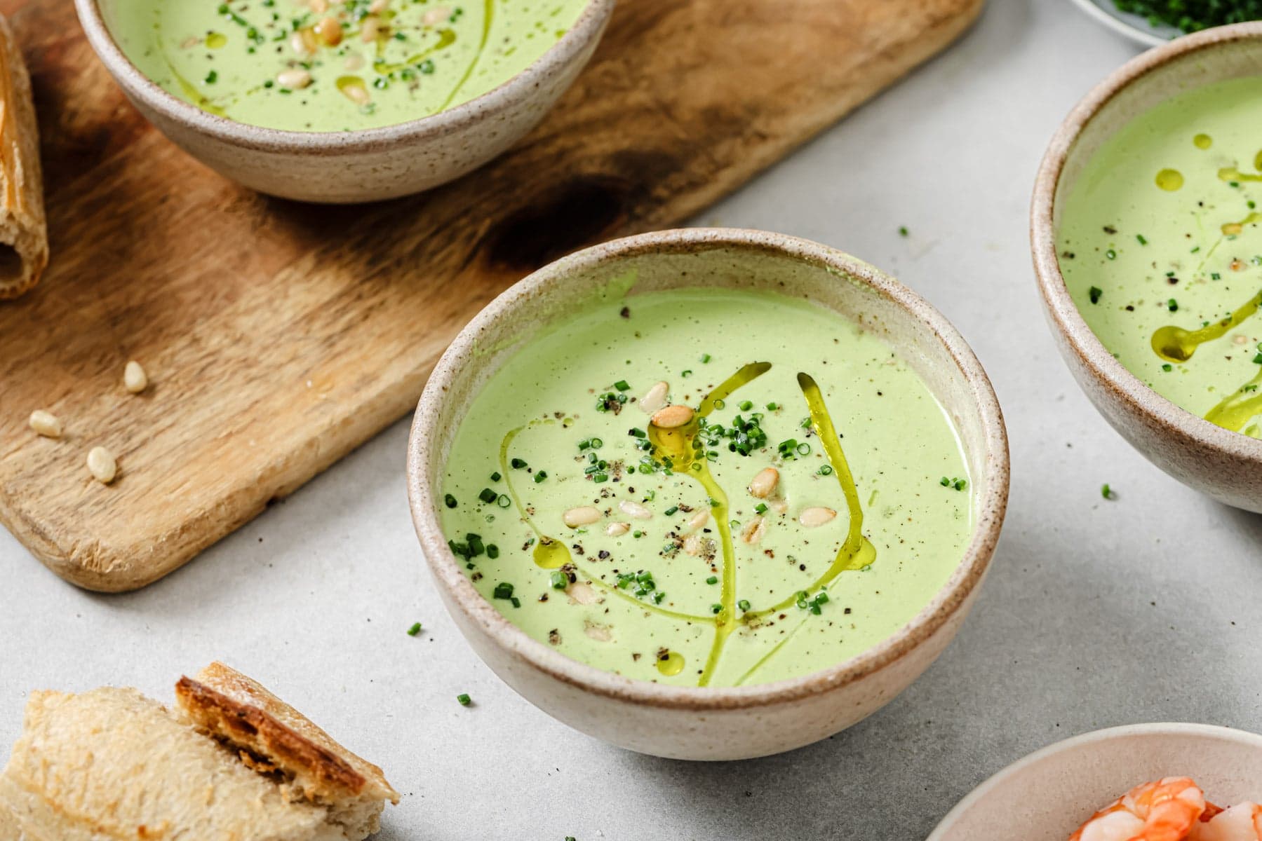 A ceramic bowl filled with creamy green soup garnished with chopped herbs, pine nuts, black pepper, and a drizzle of olive oil, with bread and more soup bowls nearby on a light surface.