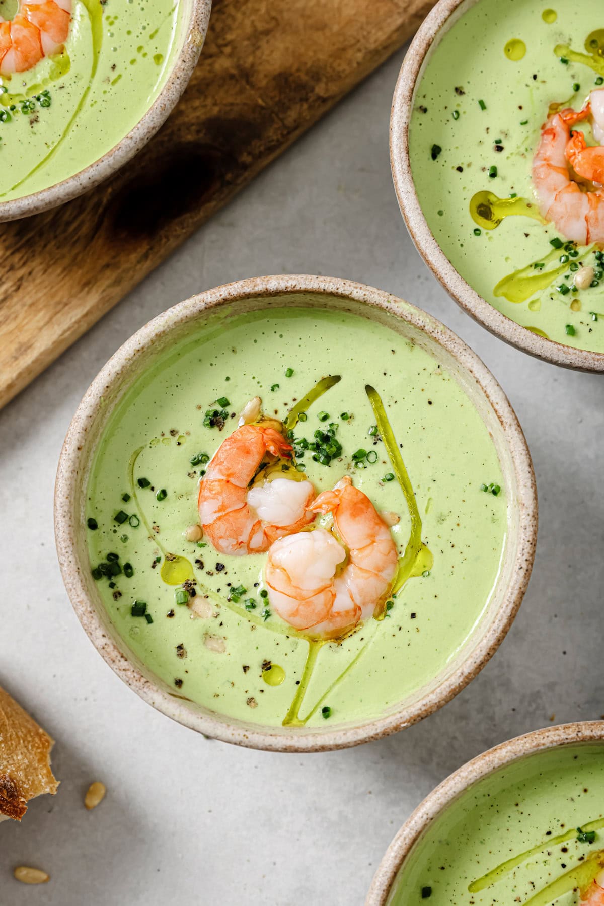 A bowl of creamy green soup garnished with two shrimp, chopped herbs, black pepper, and a drizzle of olive oil. Other similar bowls and a piece of bread are partially visible nearby.