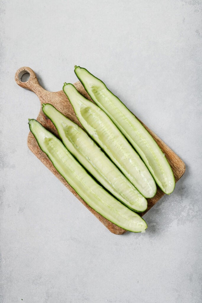 Four cucumber halves with seeds removed are placed side by side on a small wooden cutting board, on a light gray surface.