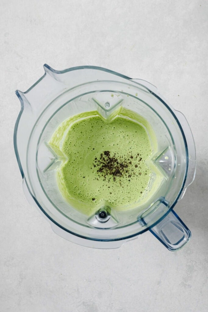 Overhead view of a blender containing a light green, frothy liquid with a small sprinkle of black pepper in the center. The blender is set on a light gray surface.