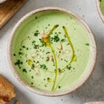 A bowl of creamy green soup garnished with a drizzle of olive oil, chopped chives, pine nuts, and ground pepper, with a piece of bread beside the bowl on a light surface.