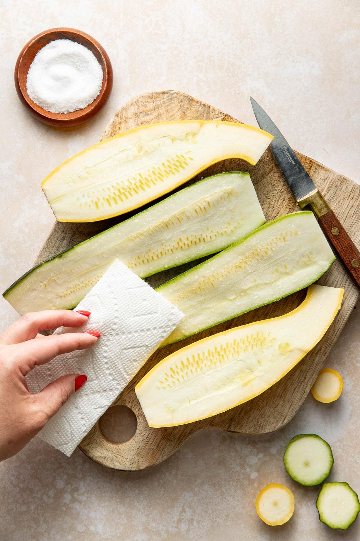A hand with red painted nails uses a paper towel to blot moisture from halved zucchini on a wooden cutting board. A knife, salt in a small bowl, and zucchini slices are nearby.
