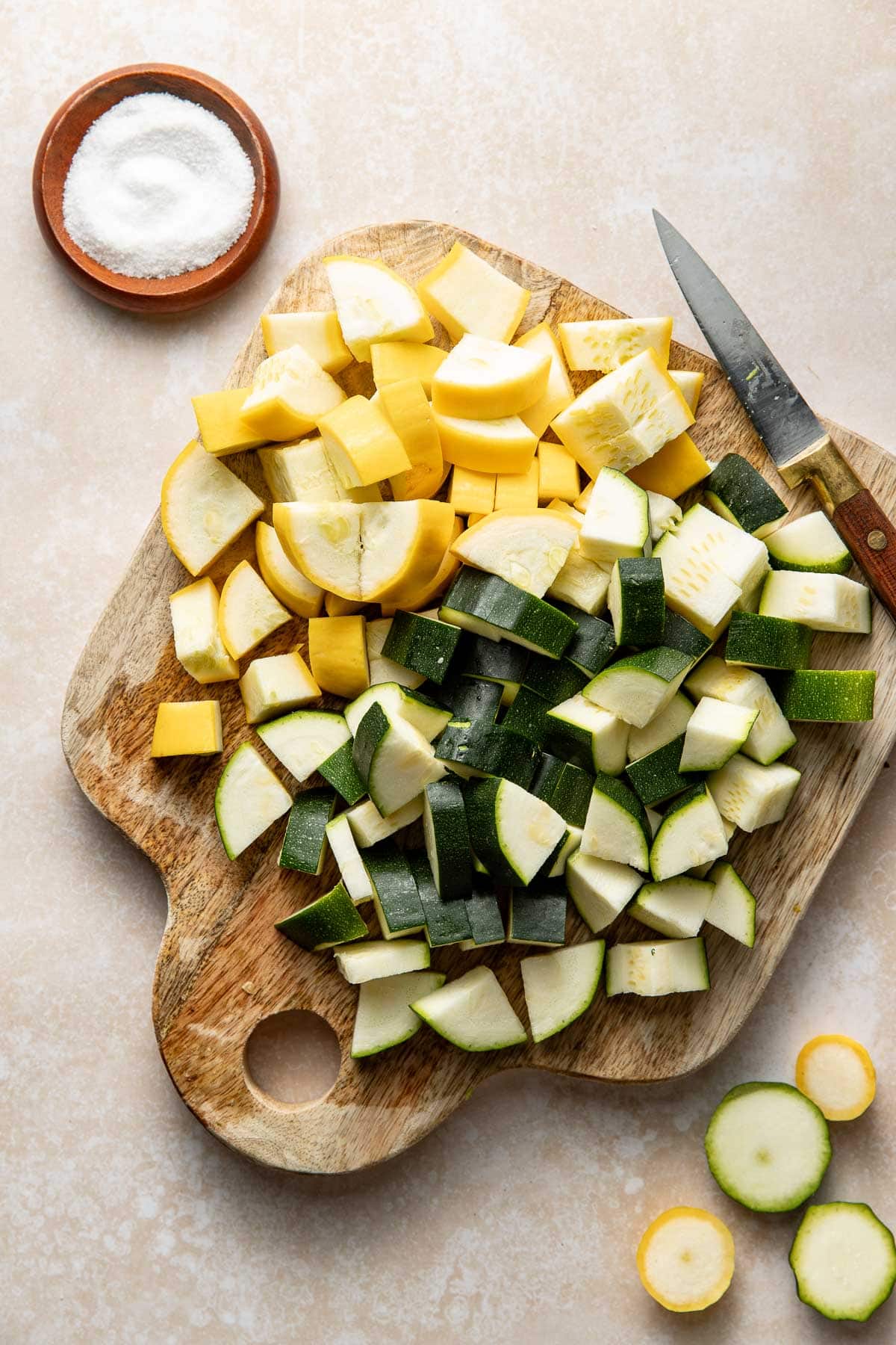 A wooden cutting board with chopped yellow squash and zucchini, a knife, and a small bowl of salt on a light countertop.