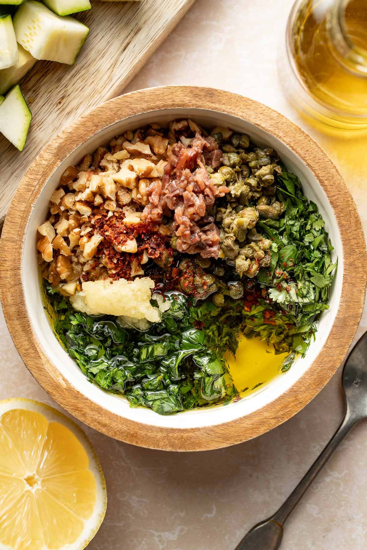 A wooden bowl filled with chopped walnuts, capers, herbs, minced garlic, olive oil, chopped anchovies, and red pepper flakes. Nearby are a lemon half, a spoon, a bottle of olive oil, and a cutting board with chopped zucchini.