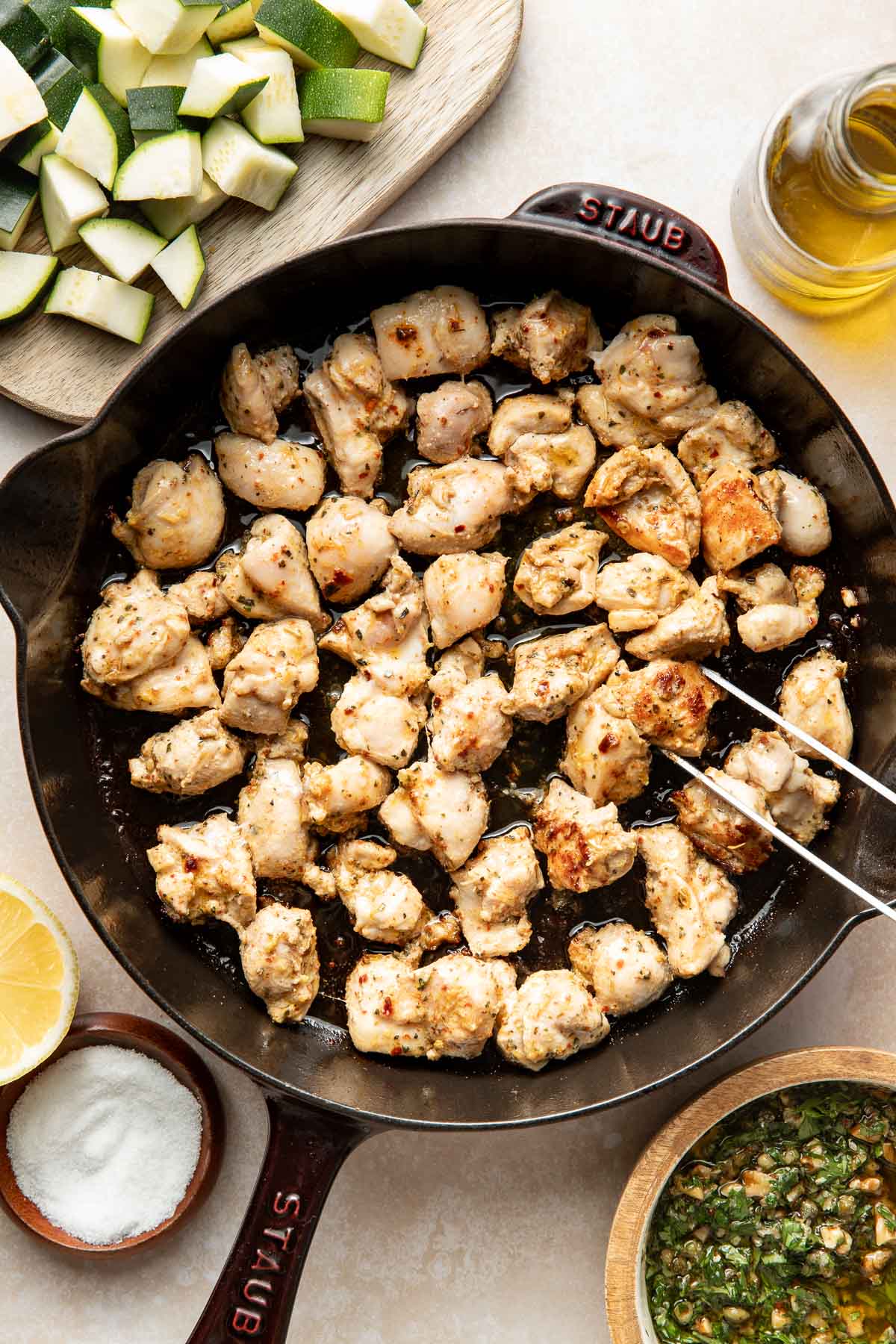 Overhead view of diced, seasoned chicken cooking in a cast iron skillet, surrounded by bowls of salt, sauce, cut zucchini, half a lemon, and a bottle of olive oil. Metal tongs are picking up a chicken piece.