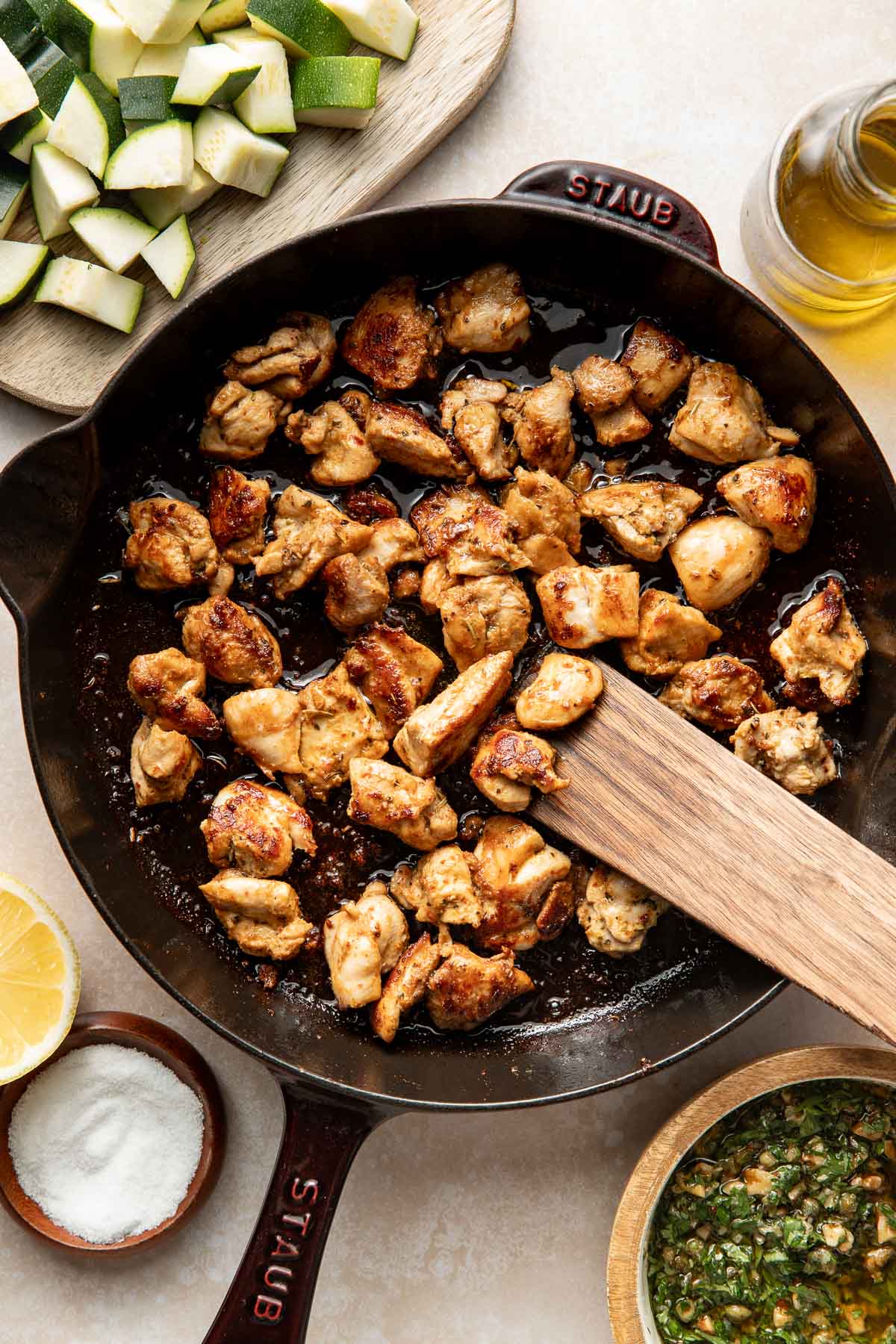 A black skillet with browned chicken pieces being stirred by a wooden spatula, surrounded by a bowl of sauce, a bowl of salt, lemon, a bottle of olive oil, and a cutting board with chopped zucchini.