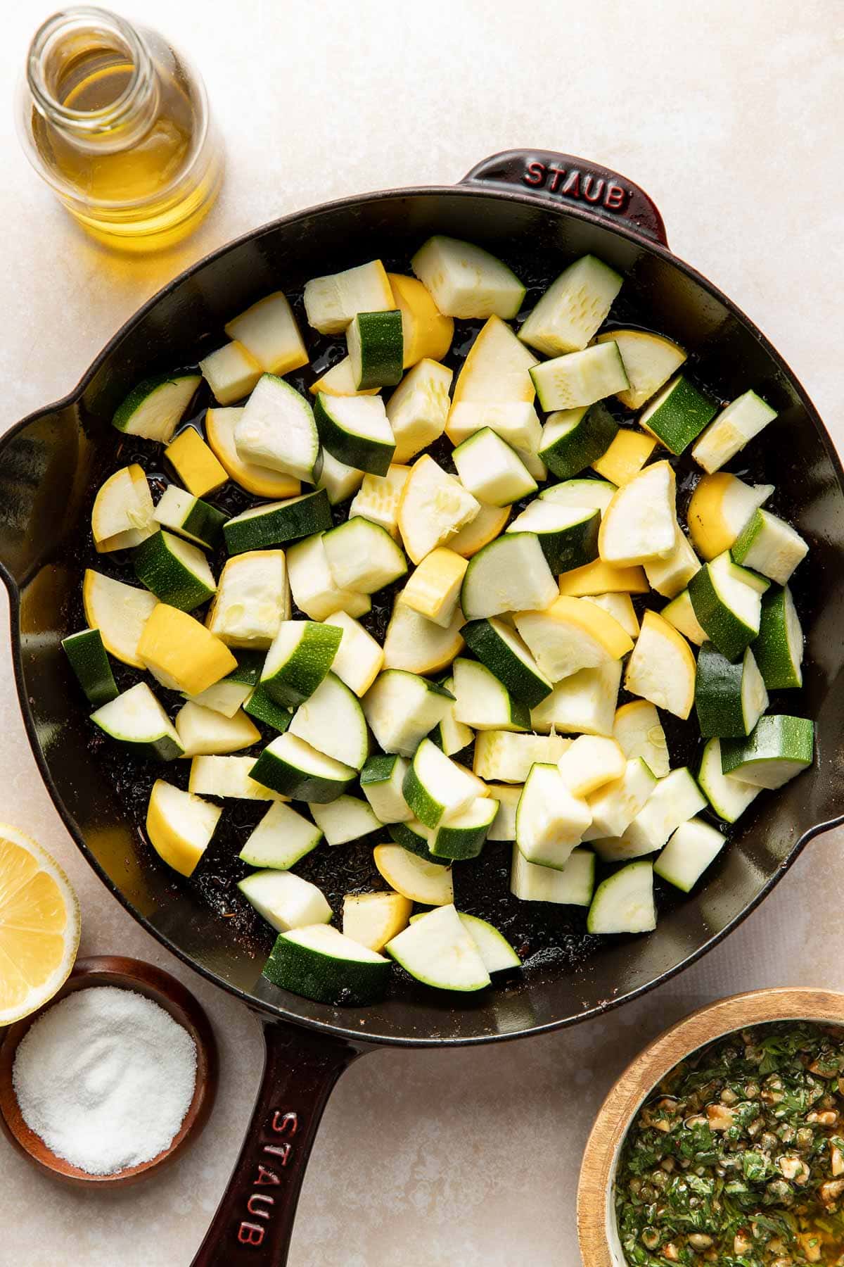 A cast iron skillet filled with chopped yellow squash and zucchini, surrounded by a bottle of olive oil, a bowl of salt, a lemon half, and a bowl of chopped herbs.