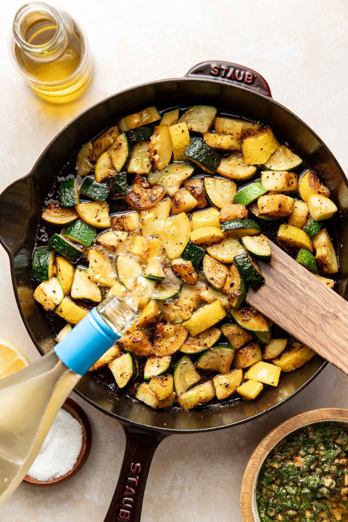 A cast iron skillet filled with sautéed zucchini and potatoes, with a wooden spatula resting inside. A hand is pouring white wine into the skillet. Nearby are a bottle of olive oil, a bowl of salt, and a bowl of sauce.