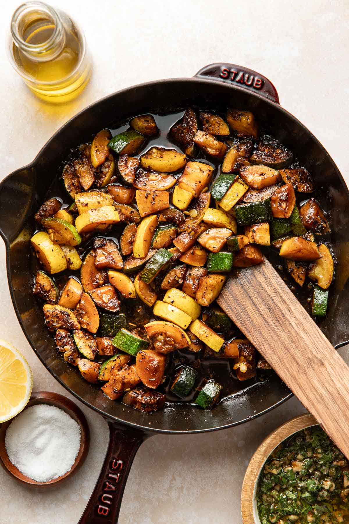 A skillet filled with sautéed mixed vegetables, including zucchini and mushrooms, sits on a light surface. Surrounding the pan are a small bowl of salt, half a lemon, a bottle of oil, and a bowl of green sauce.