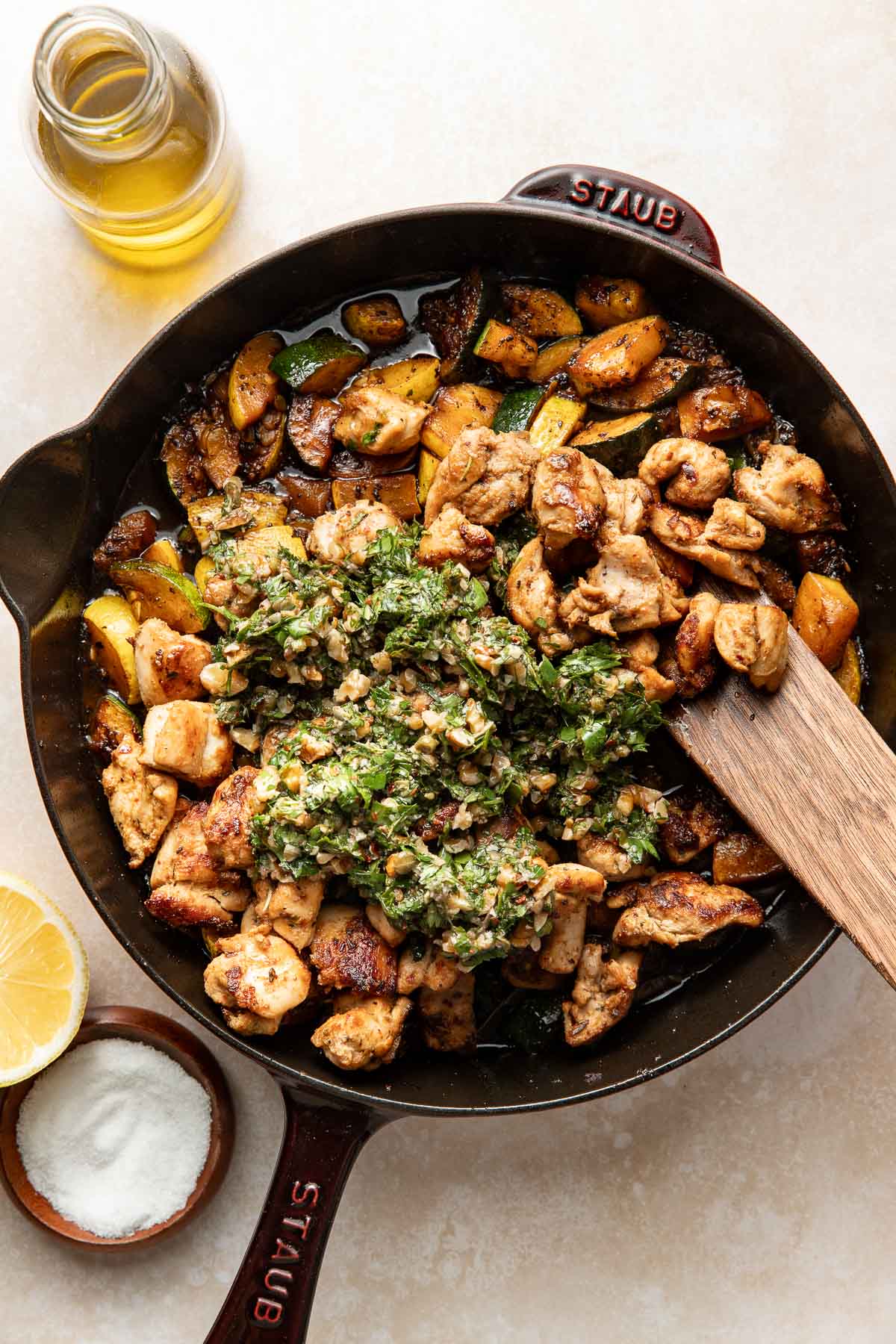 A cast iron skillet filled with cooked chicken, sautéed vegetables, and topped with a green herb sauce. Nearby are a wooden spoon, a bottle of olive oil, a bowl of salt, and a lemon half.