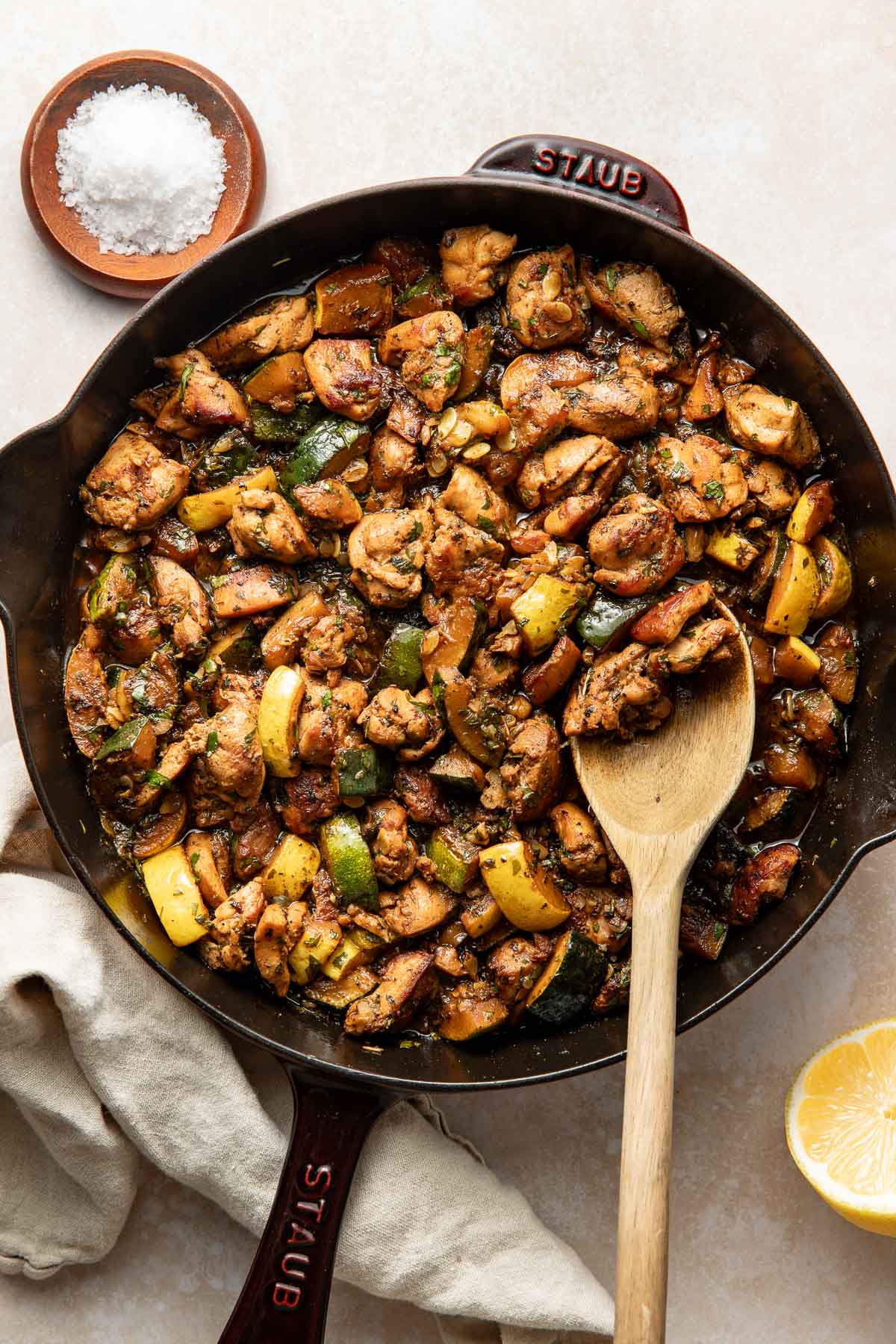 A large skillet filled with sautéed chicken pieces, chopped green and yellow vegetables, and herbs, with a wooden spoon resting inside. A bowl of salt, a lemon half, and a beige napkin are placed nearby.